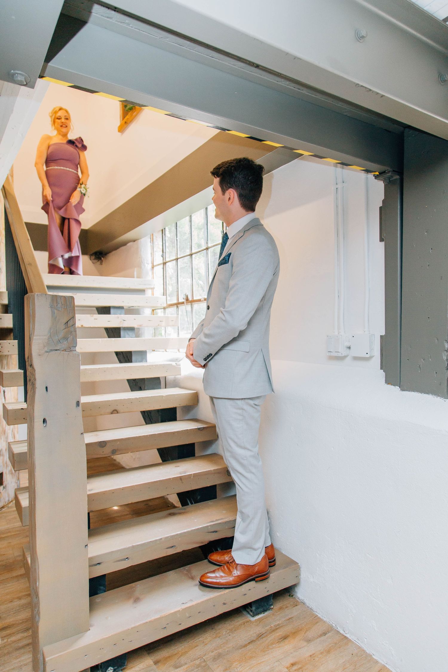 Groom in gray suit stands on modern wooden staircase looking up at bridesmaid in lavender dress above.