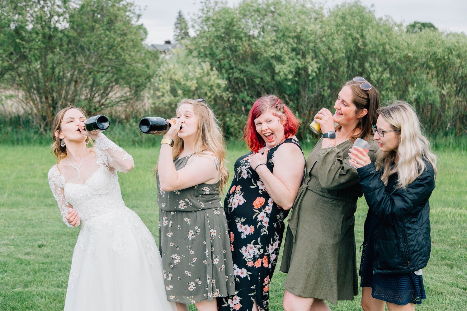 Bride and bridesmaids drinking champagne and laughing together outdoors after wedding ceremony.
