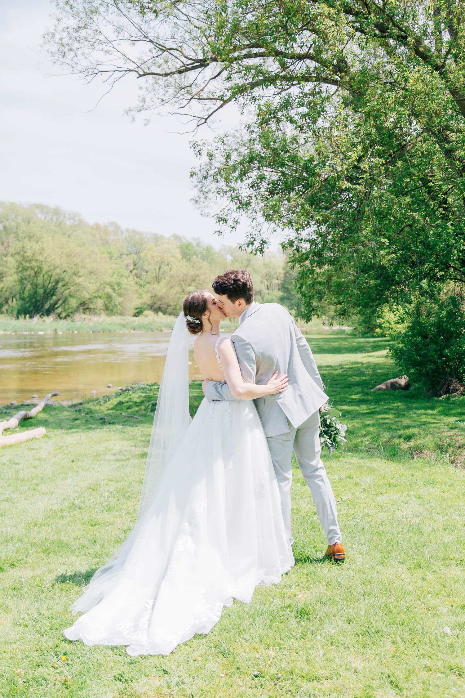 Bride and groom share a kiss beside a river surrounded by lush green trees.