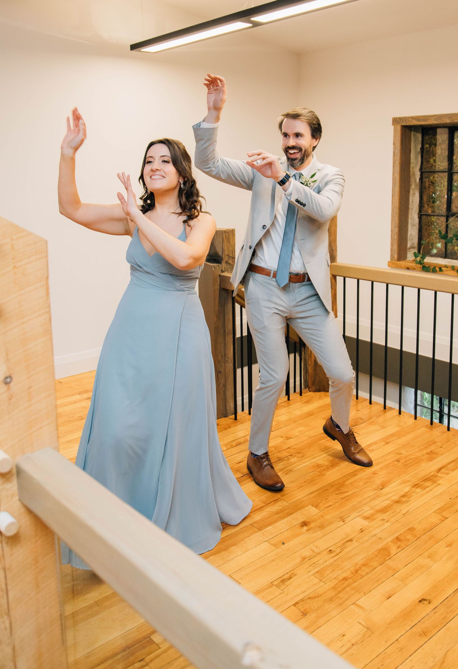 Bridesmaid in blue dress and groomsman in gray suit dancing with raised arms at wedding reception.