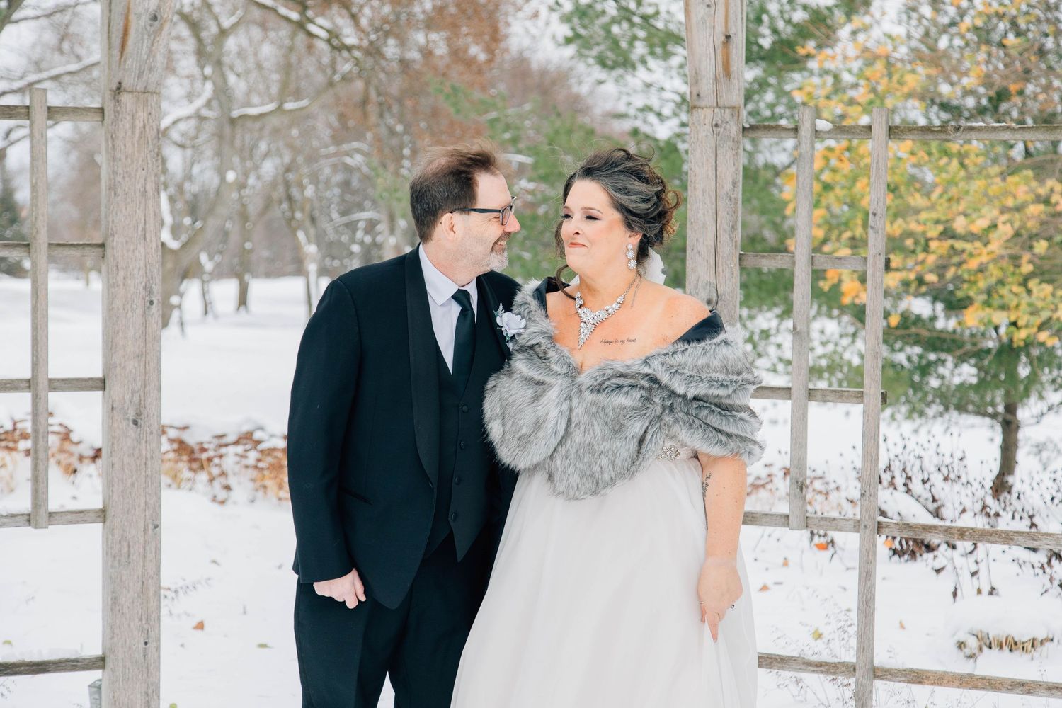 Bride and groom share a romantic glance under a rustic wooden arbor in a snowy winter setting.