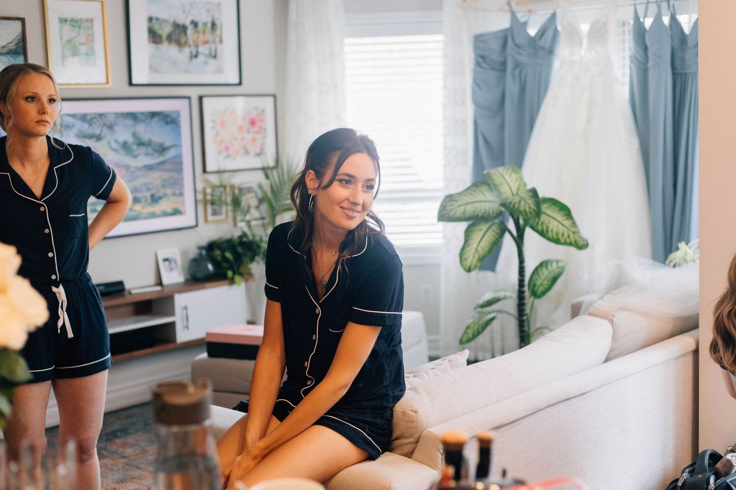 Bridesmaid in navy pajamas sits smiling with wedding dress visible in background.