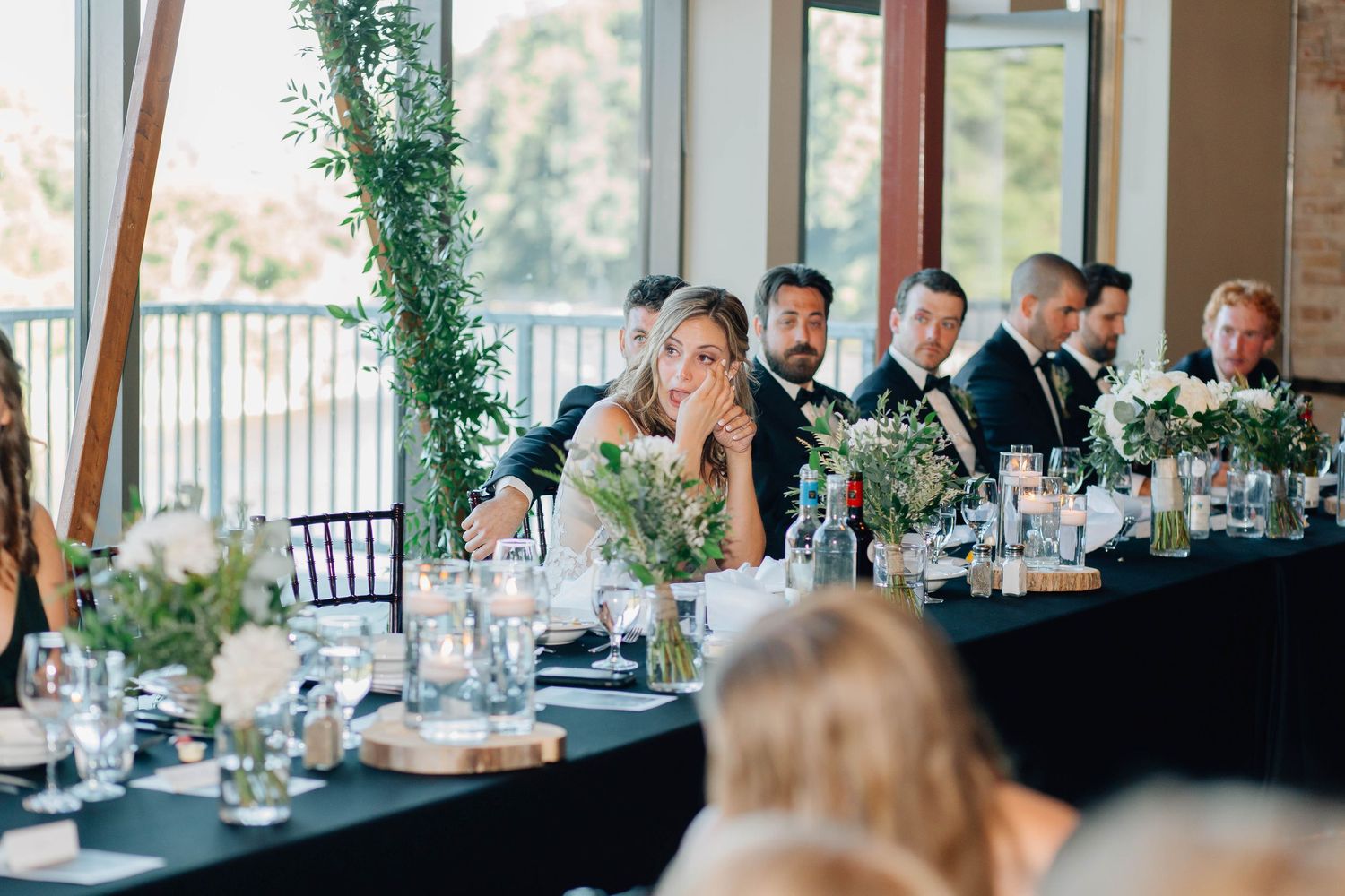 Wedding party seated at a long head table with lush greenery centerpieces during elegant indoor reception.