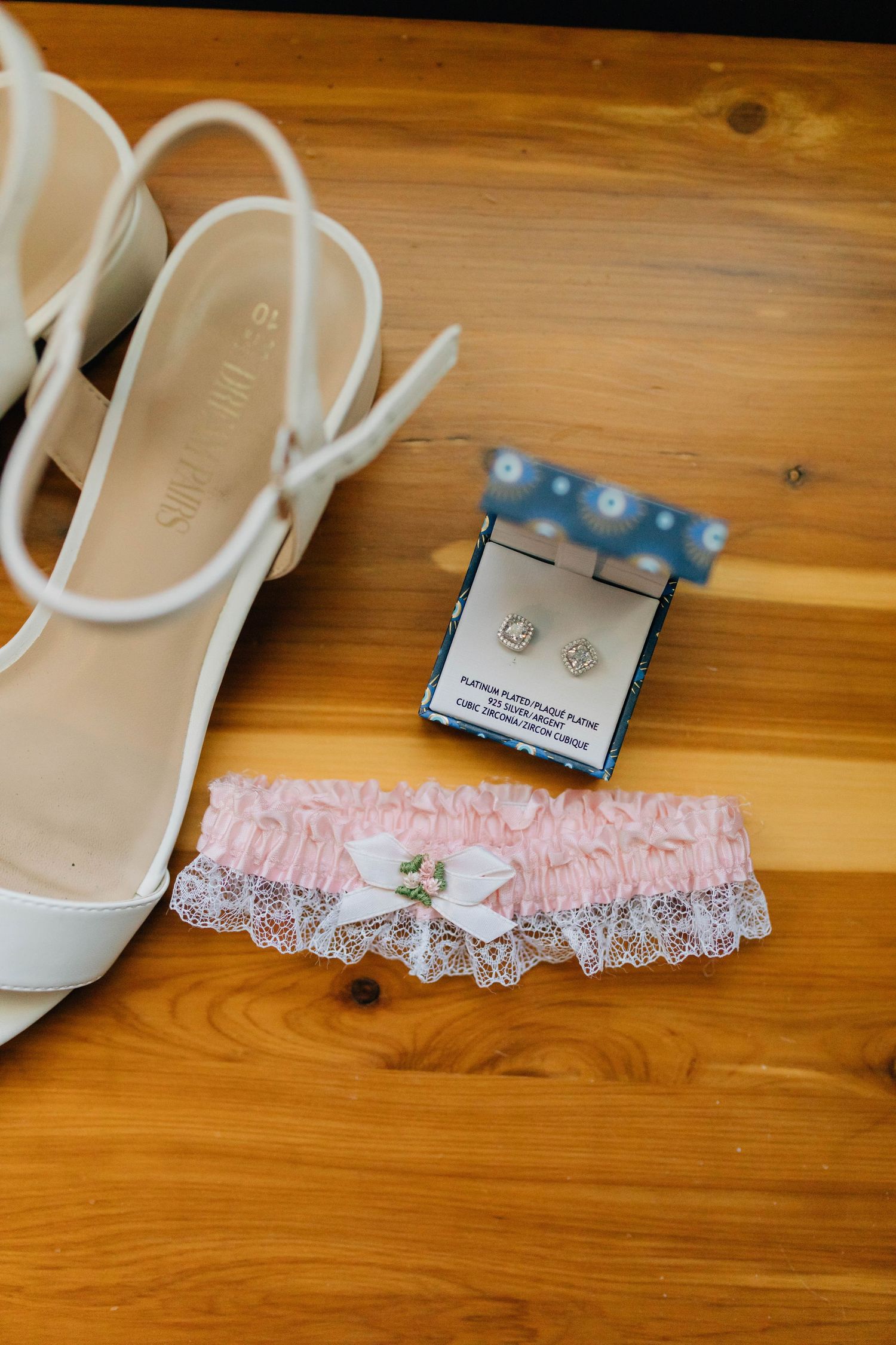 White bridal heels, pink lace garter, and diamond earrings in a blue gift box arranged on a wooden surface.