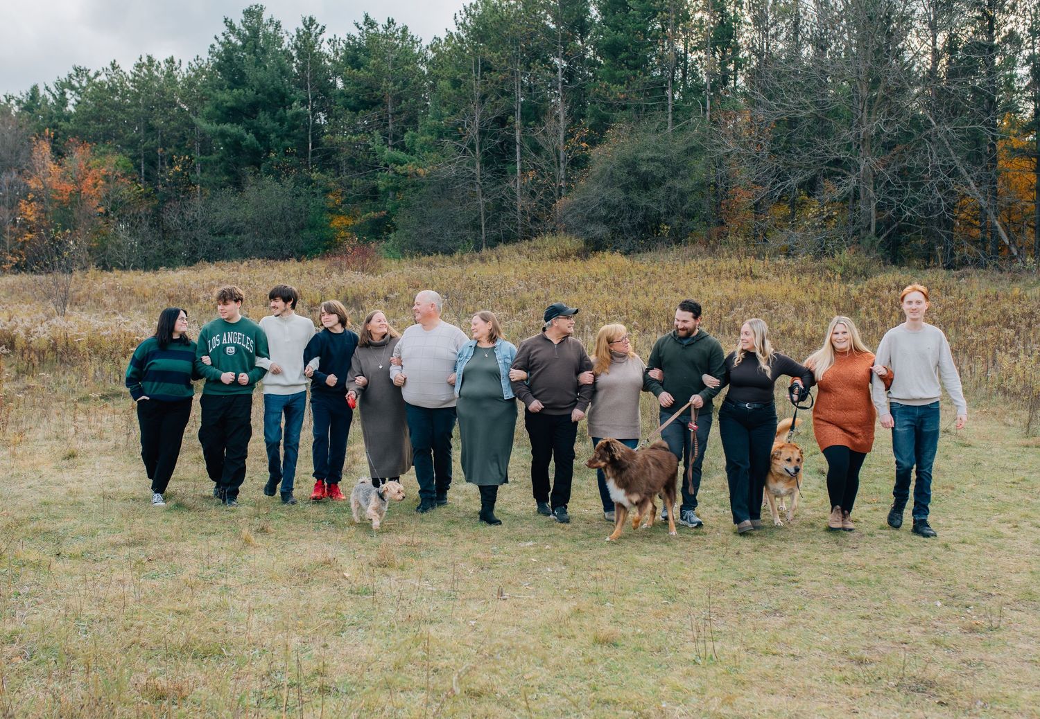 Large extended family walking together with dogs through an open field with autumn trees in background.