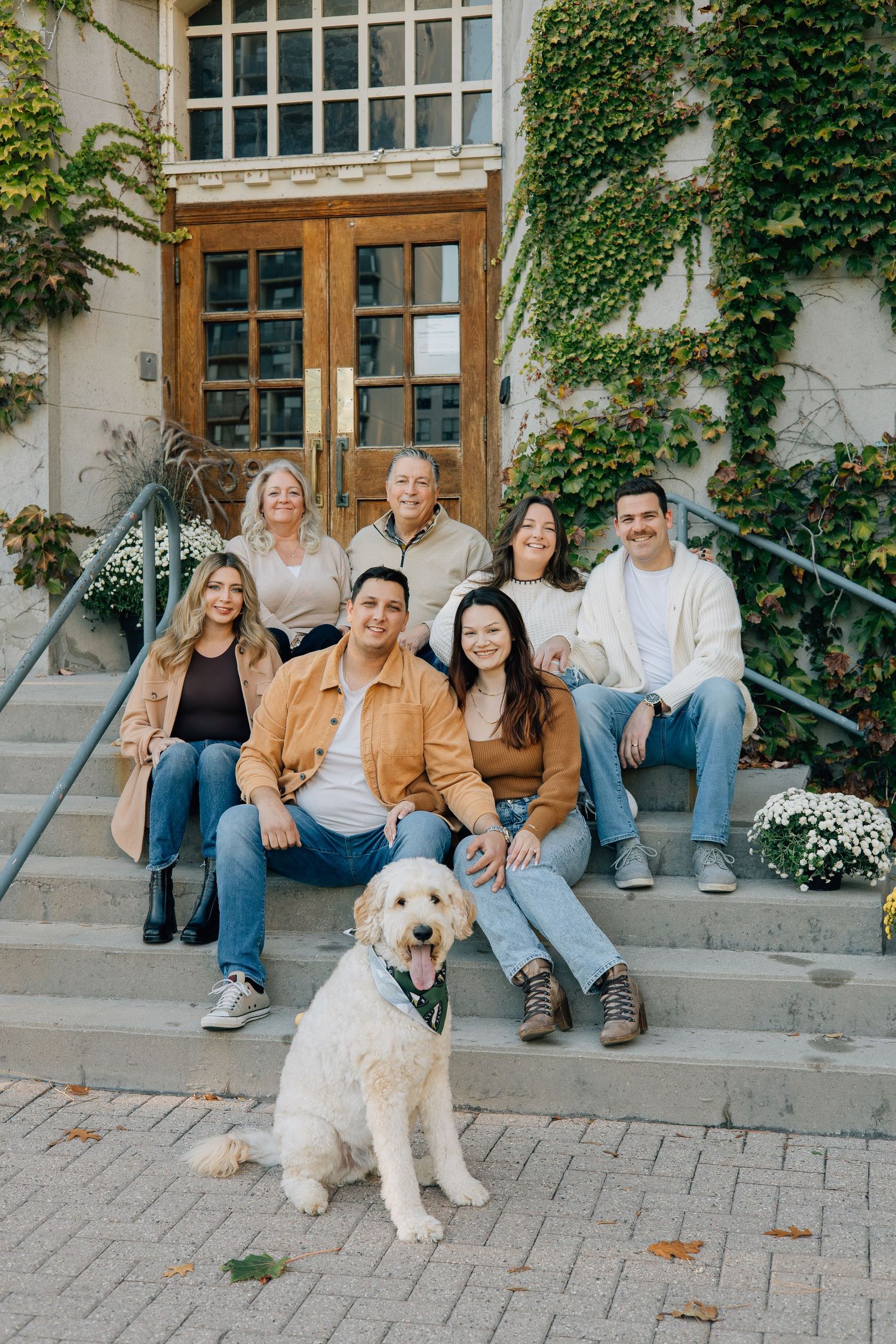 Extended family of six posing on stone steps with a white goldendoodle dog outside a building.