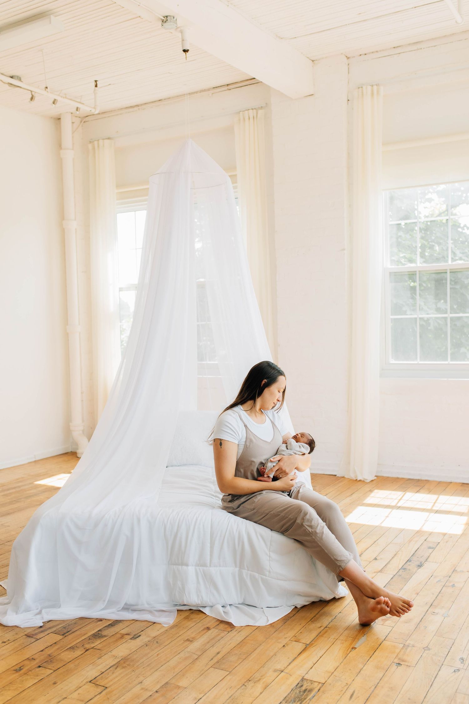 Asian mother sitting under white canopy on sunlit hardwood floor cradling sleeping newborn baby.