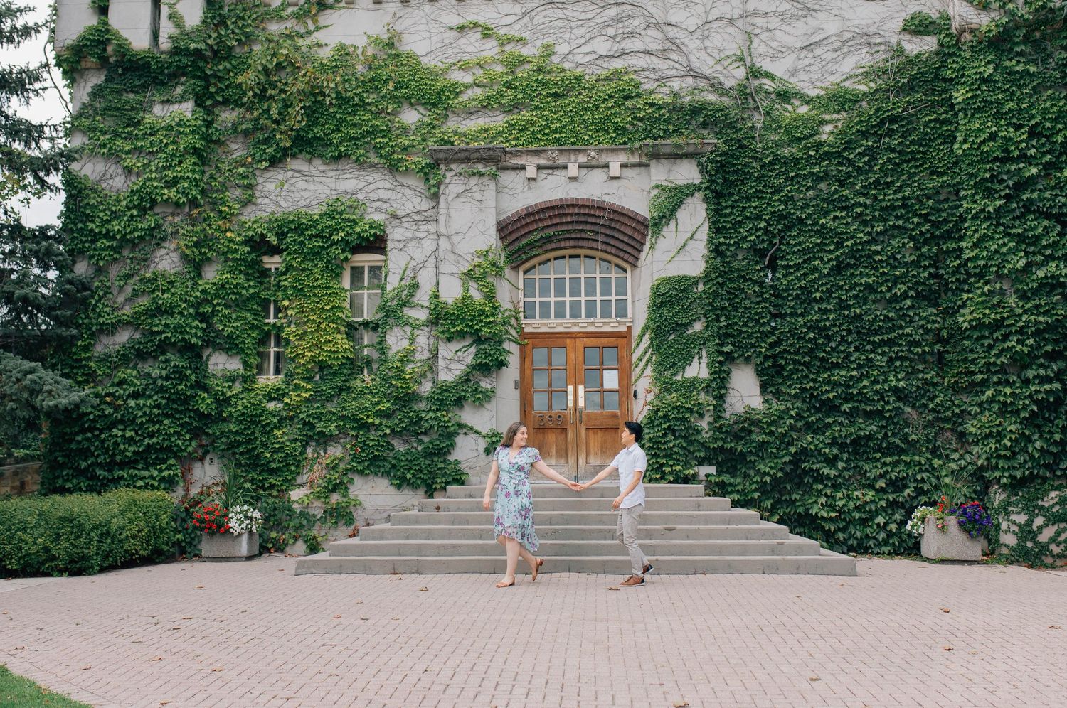 Couple holding hands walking in front of steps toward ivy-covered stone building with arched wooden doors.
