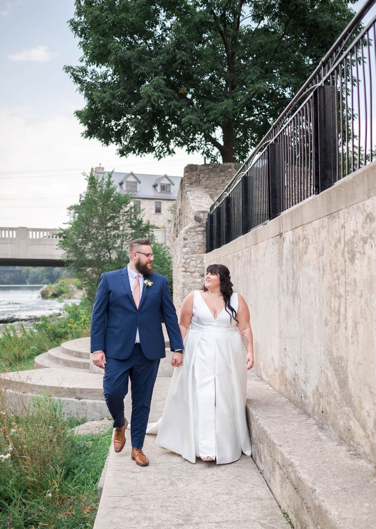 Bride and groom walk hand in hand along a scenic riverside path, bride in white gown, groom in navy suit.