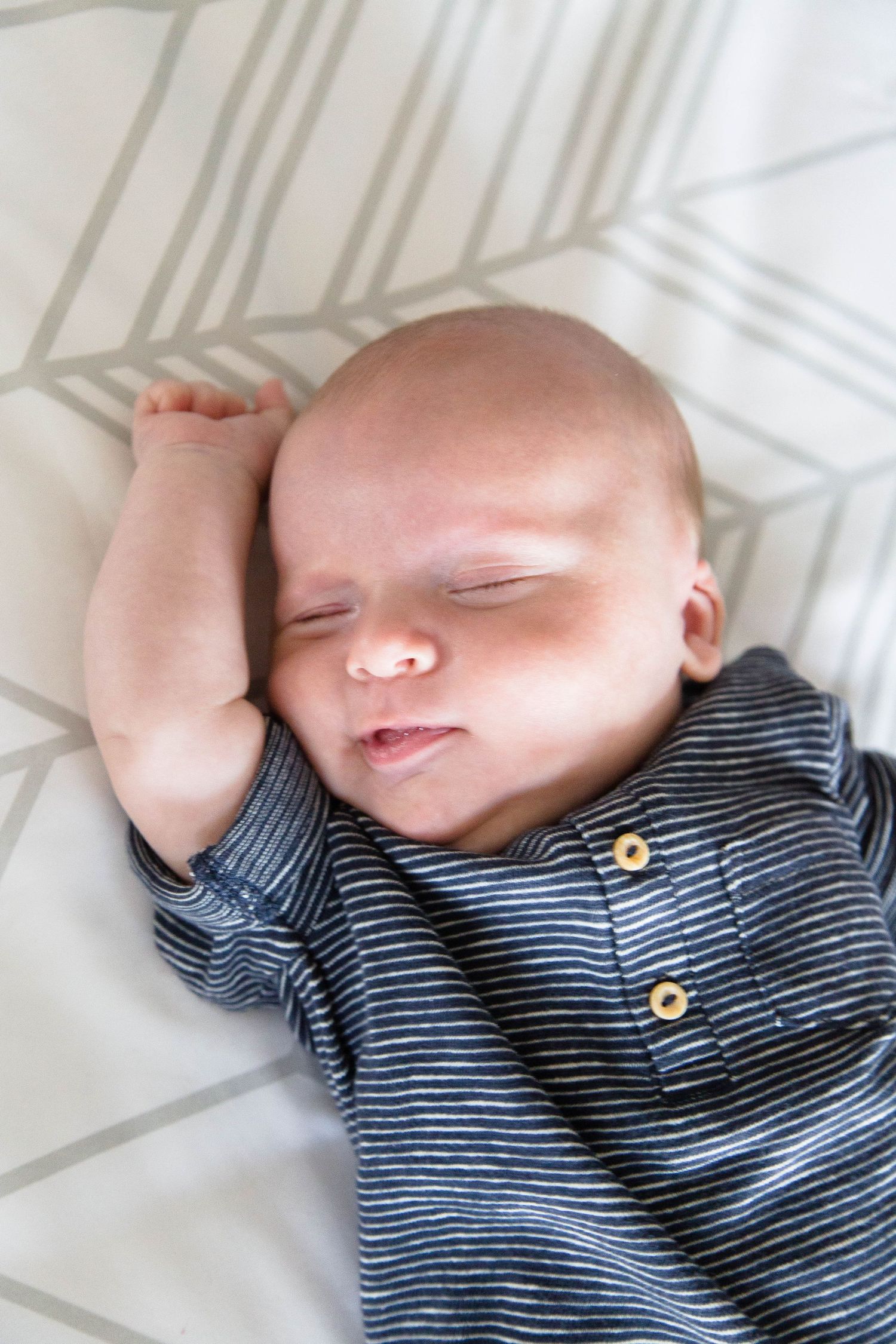 Sleeping newborn baby in striped onesie with arm raised on white bedding.
