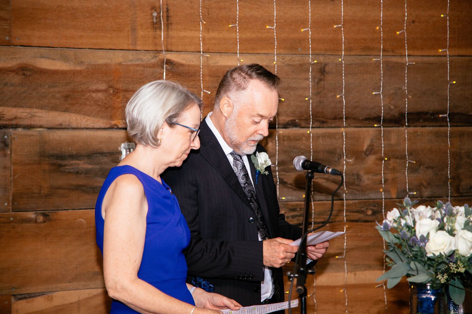 Couple in formal attire reading heartfelt speech at microphone during rustic barn wedding reception.