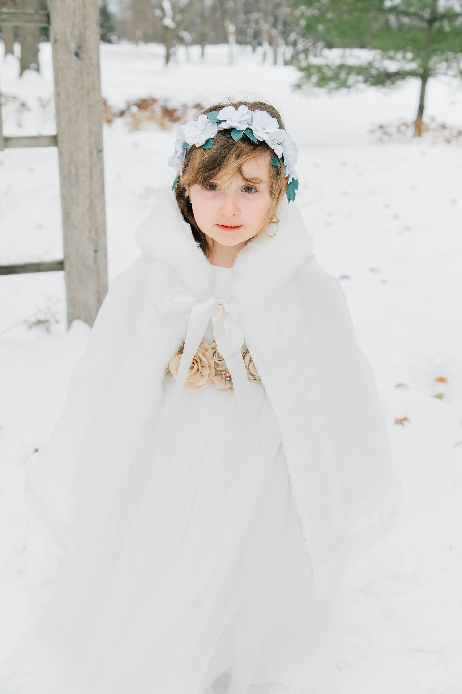 Young flower girl in white fur cape and floral crown poses in snowy winter wedding setting.