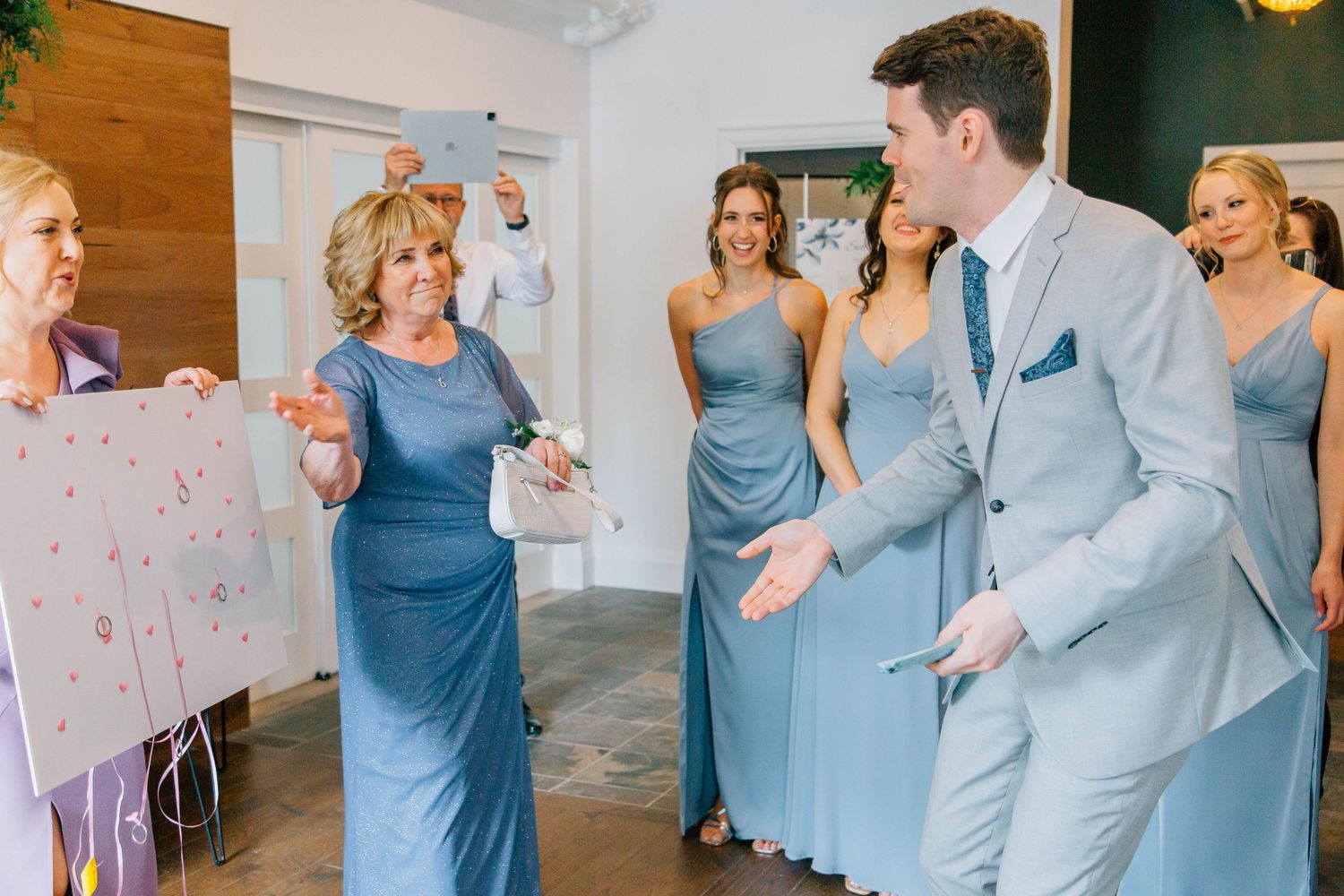 Groom gestures excitedly at wedding poster held by older woman in blue gown surrounded by bridesmaids.