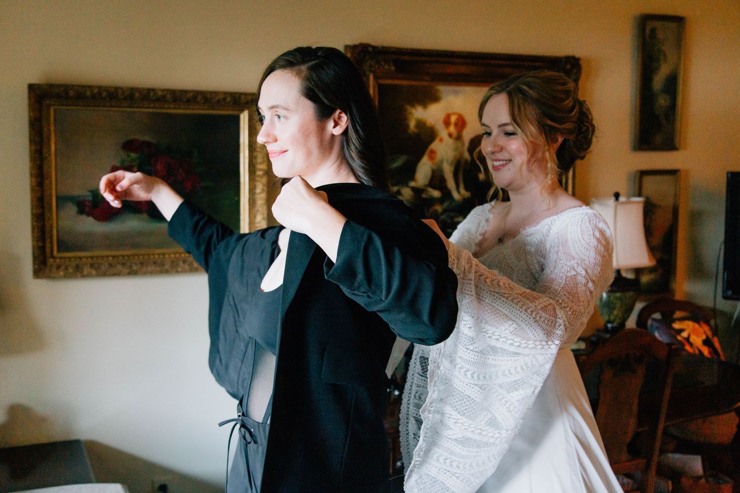 Bride in white lace gown helps partner in black blazer get dressed before wedding ceremony.