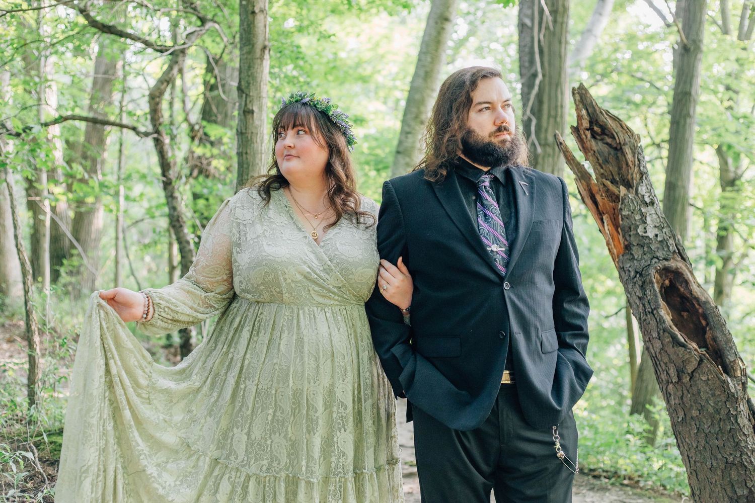 Bride in sage green lace gown and groom in black suit posing in a lush forest setting.
