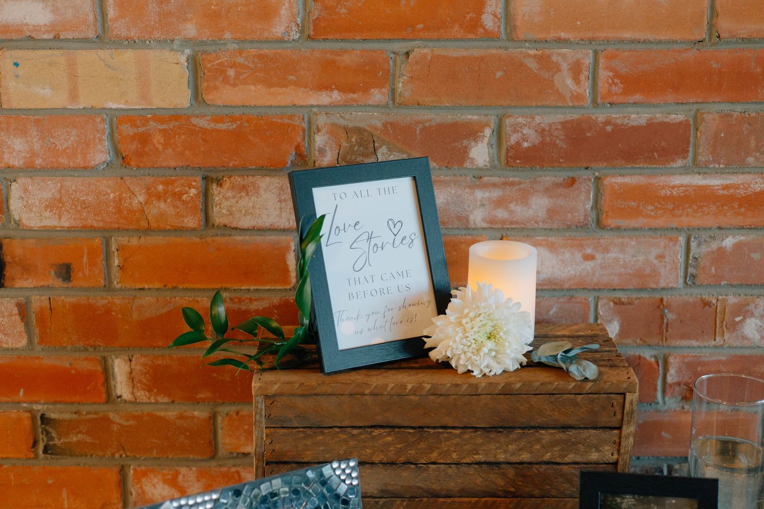 A framed wedding welcome sign with a white flower and candle displayed against a brick wall.