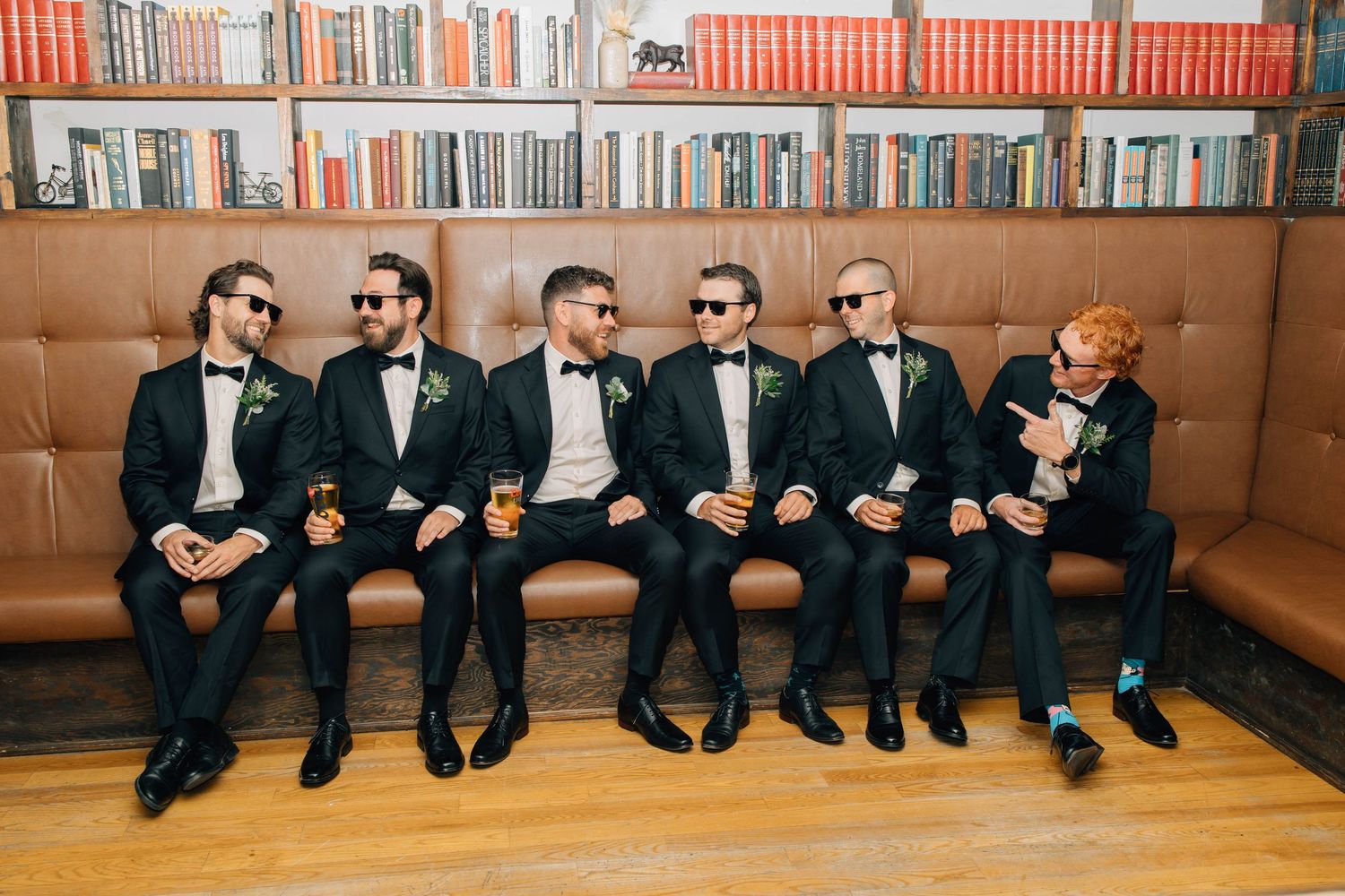 Six groomsmen in black tuxedos wearing sunglasses pose on a leather sofa in front of bookshelves.