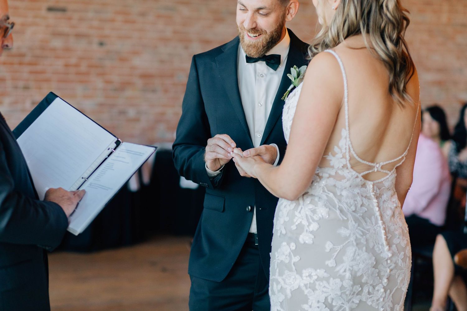 Groom placing wedding ring on brides finger during indoor ceremony with officiant.