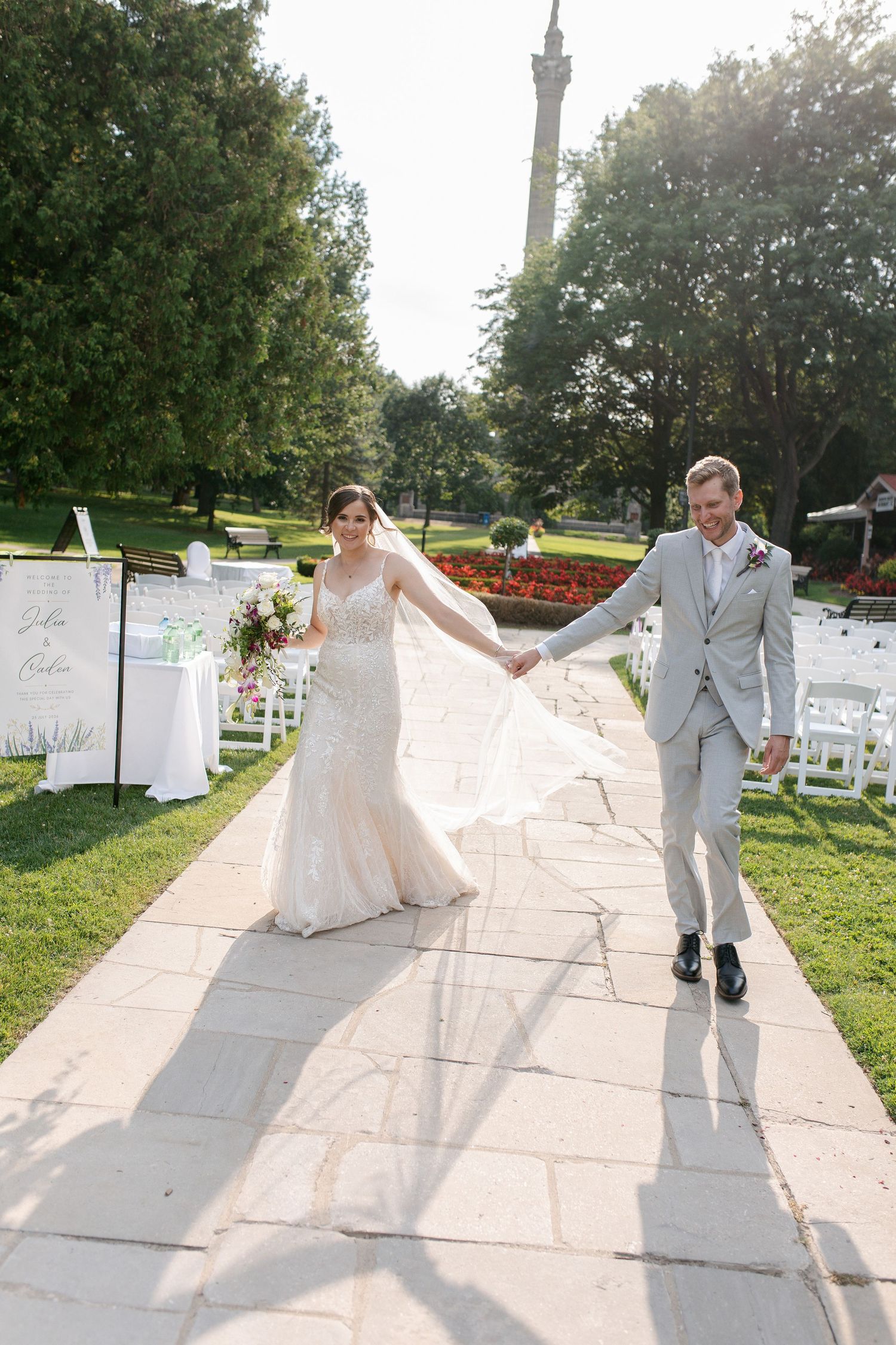 Bride and groom walk hand-in-hand down outdoor wedding aisle, bride in lace gown with flowing veil, groom in gray suit just after ceremony.