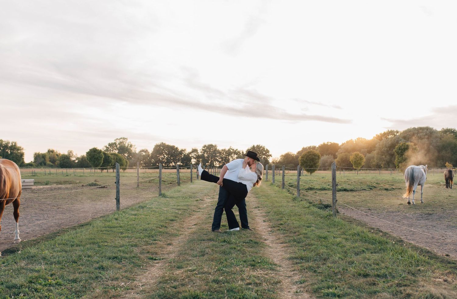 A couple sharing a romantic dip embrace on a grassy farm path at sunset with horses nearby.