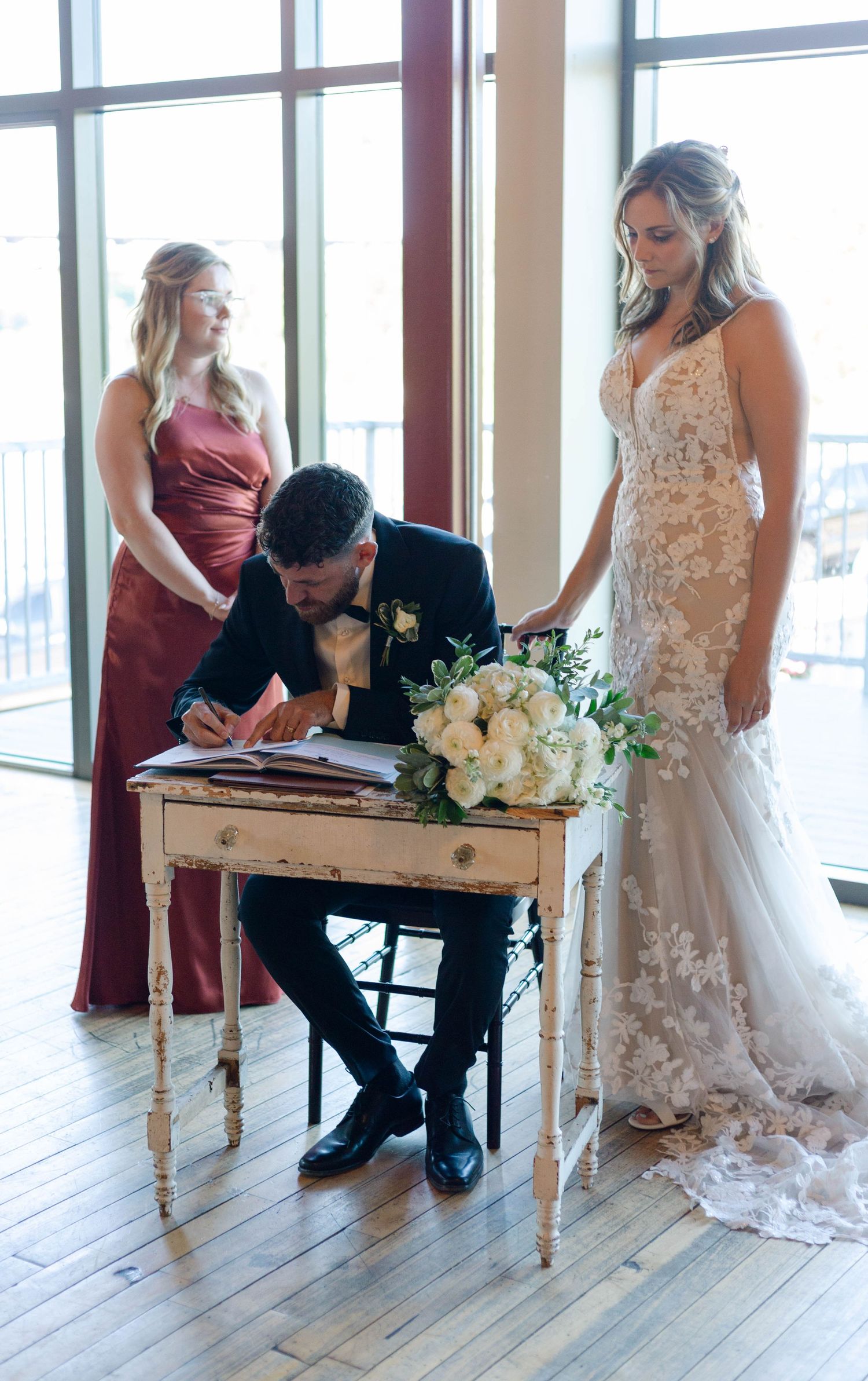 Groom signing marriage certificate at vintage desk with bride in lace gown and bridesmaid in burgundy dress.