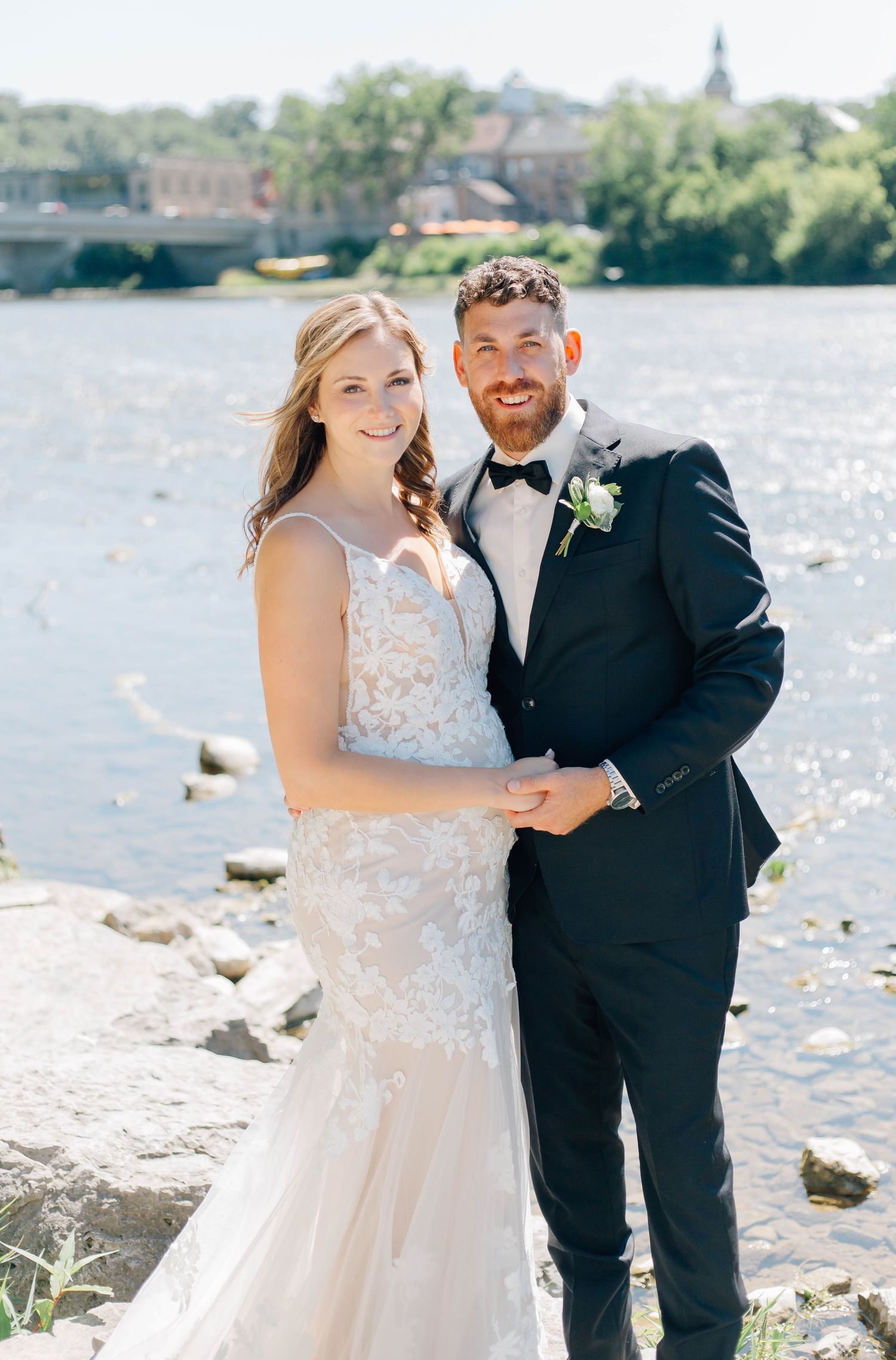 Bride and groom smiling together by a riverside on their wedding day, wearing a lace gown and navy tuxedo.