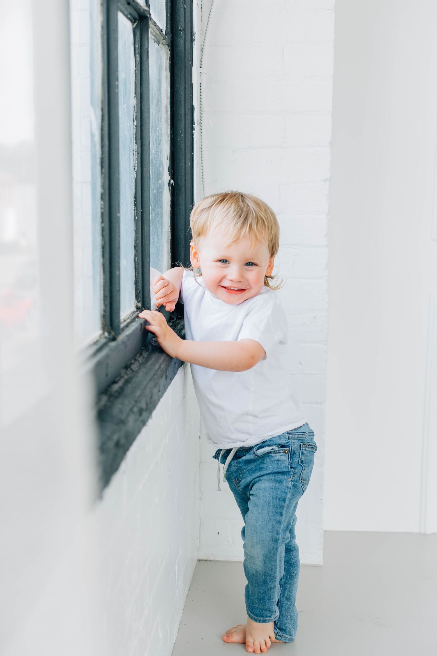 Happy toddler boy in white tee and jeans standing by black-framed window on white brick wall.