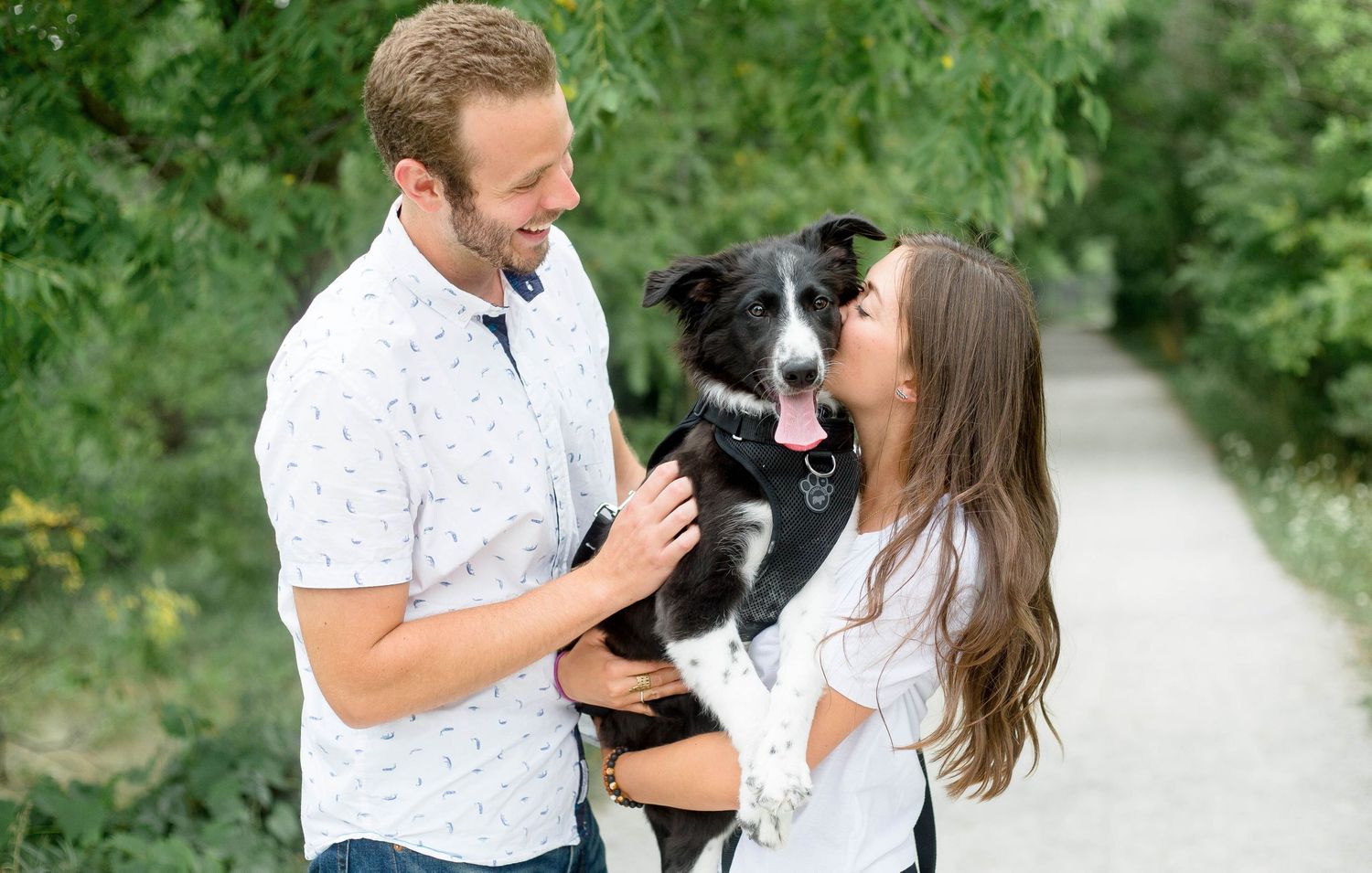 Couple smiling at their black and white Border Collie dog while holding it on a tree-lined gravel path.
