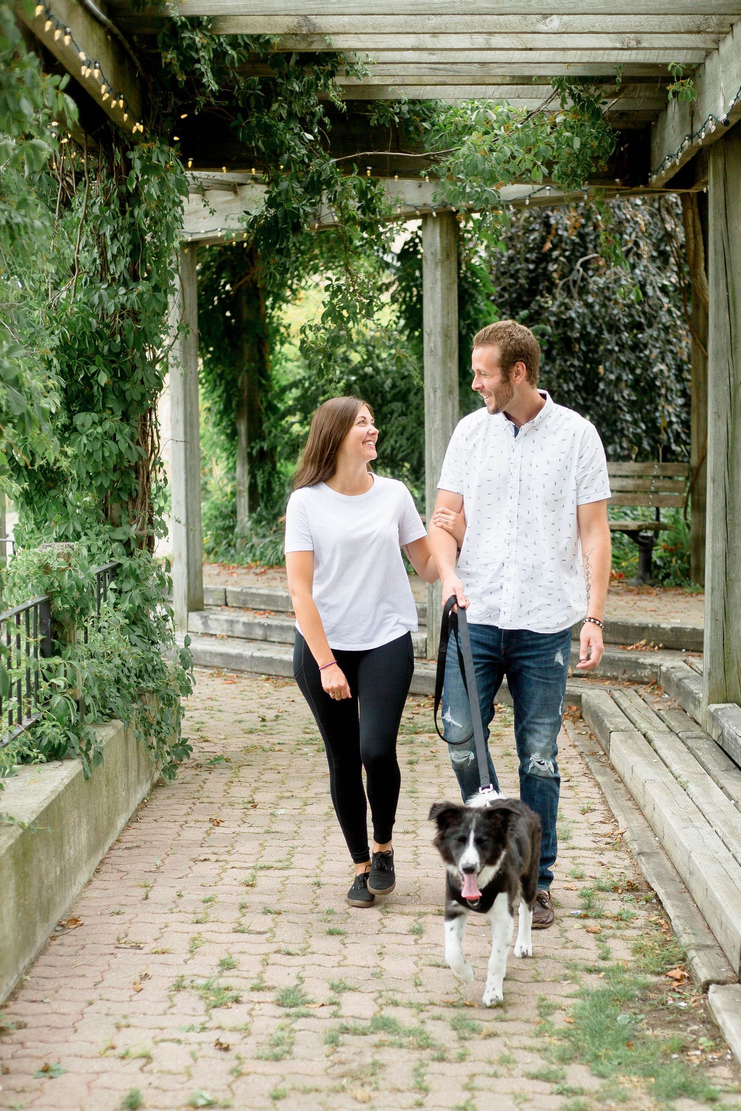A couple walks hand in hand with their Border Collie through a lush garden pergola pathway.
