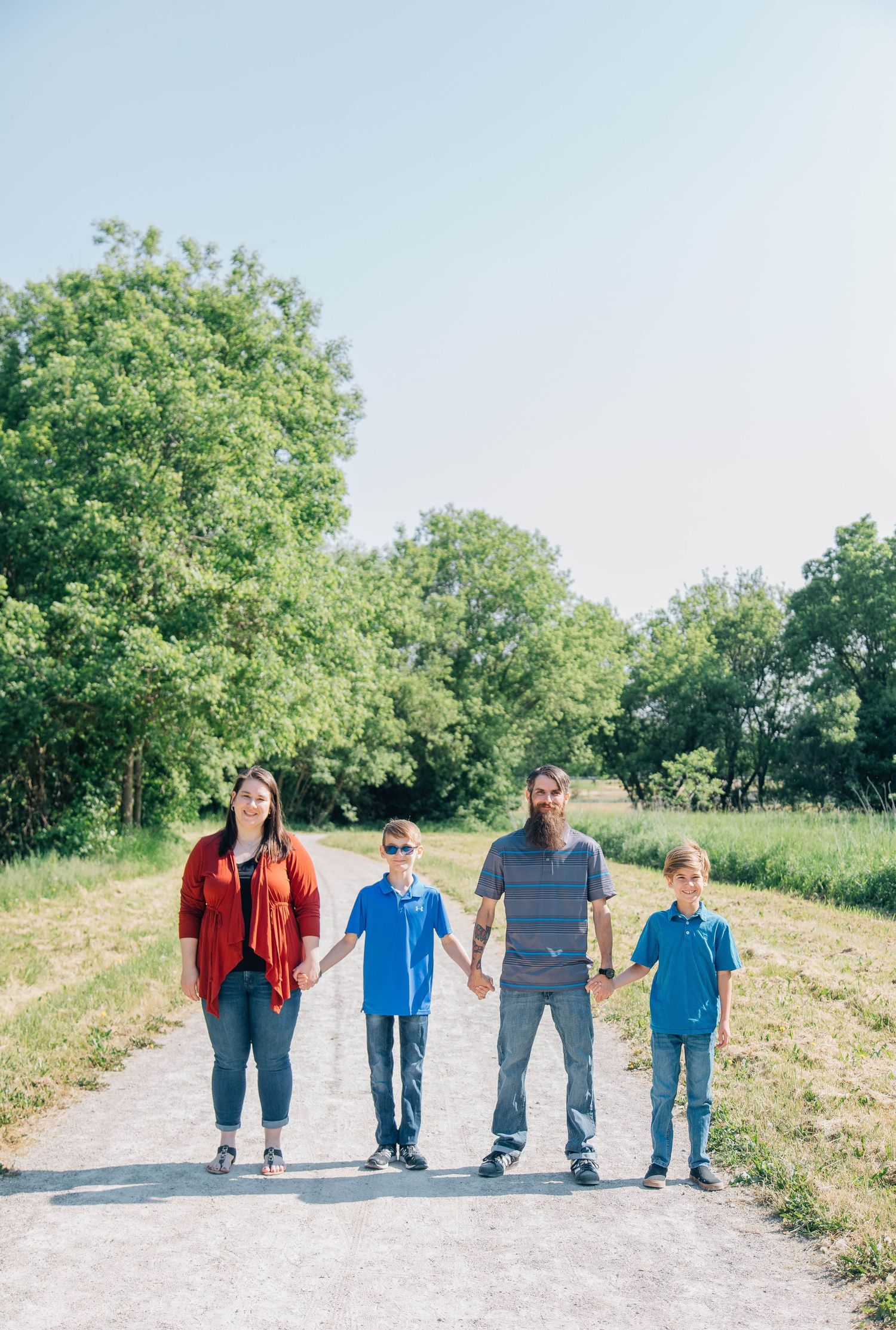 Family of four holding hands on a sunny gravel path surrounded by green trees.
