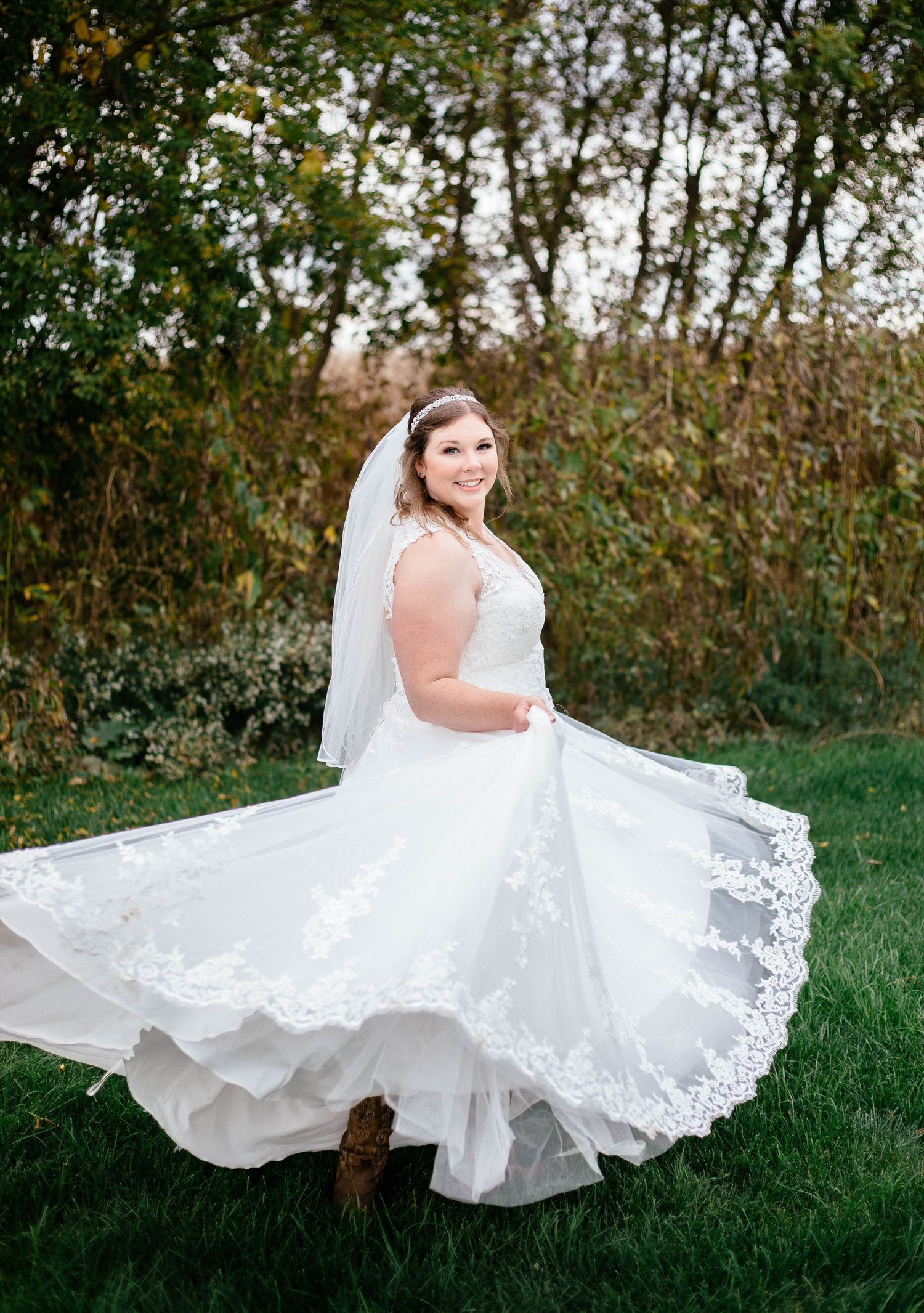 Bride twirls in her lace wedding gown and veil on a green lawn with autumn foliage backdrop.
