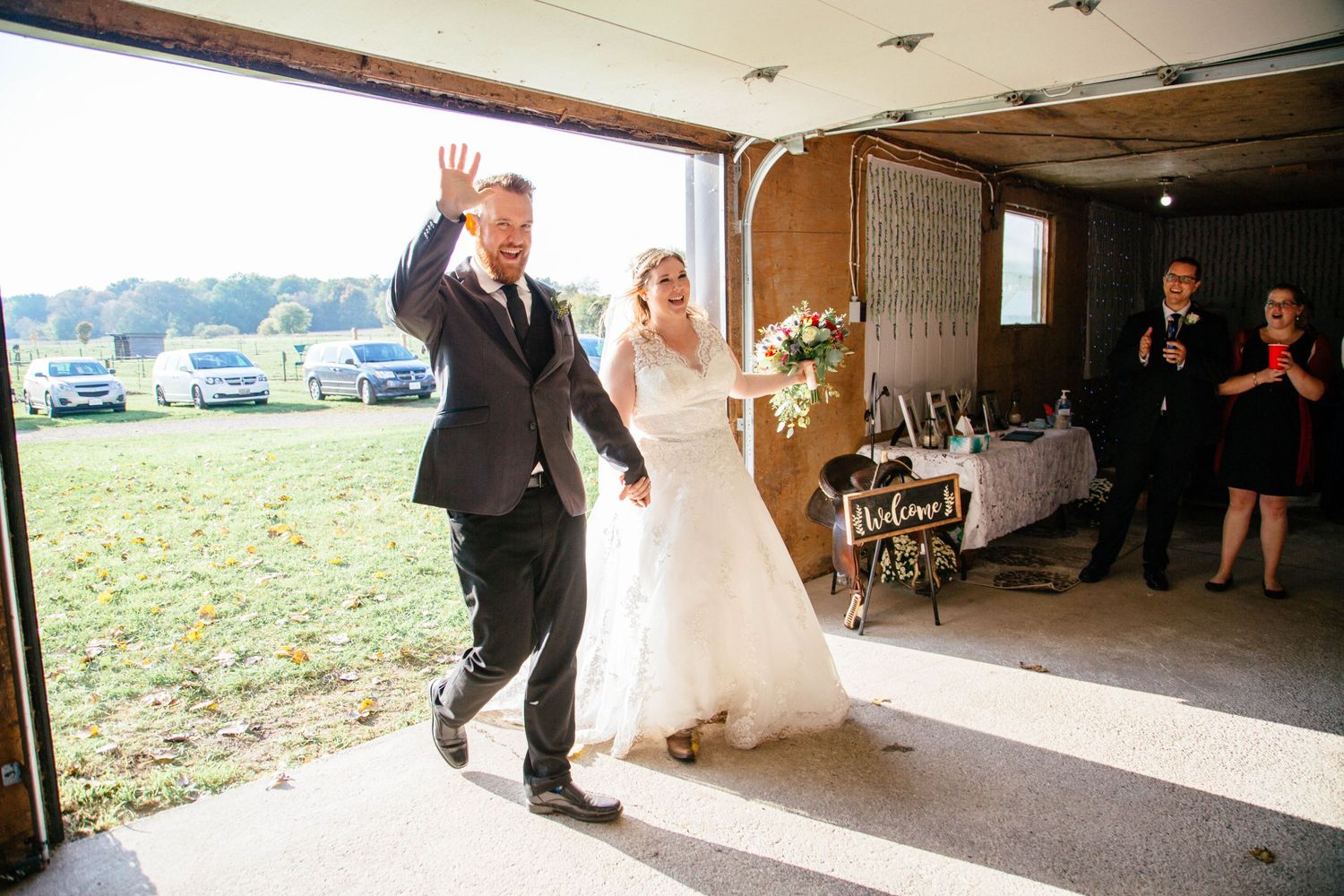 Bride and groom make grand entrance into rustic barn wedding reception as groom waves to seated guests.