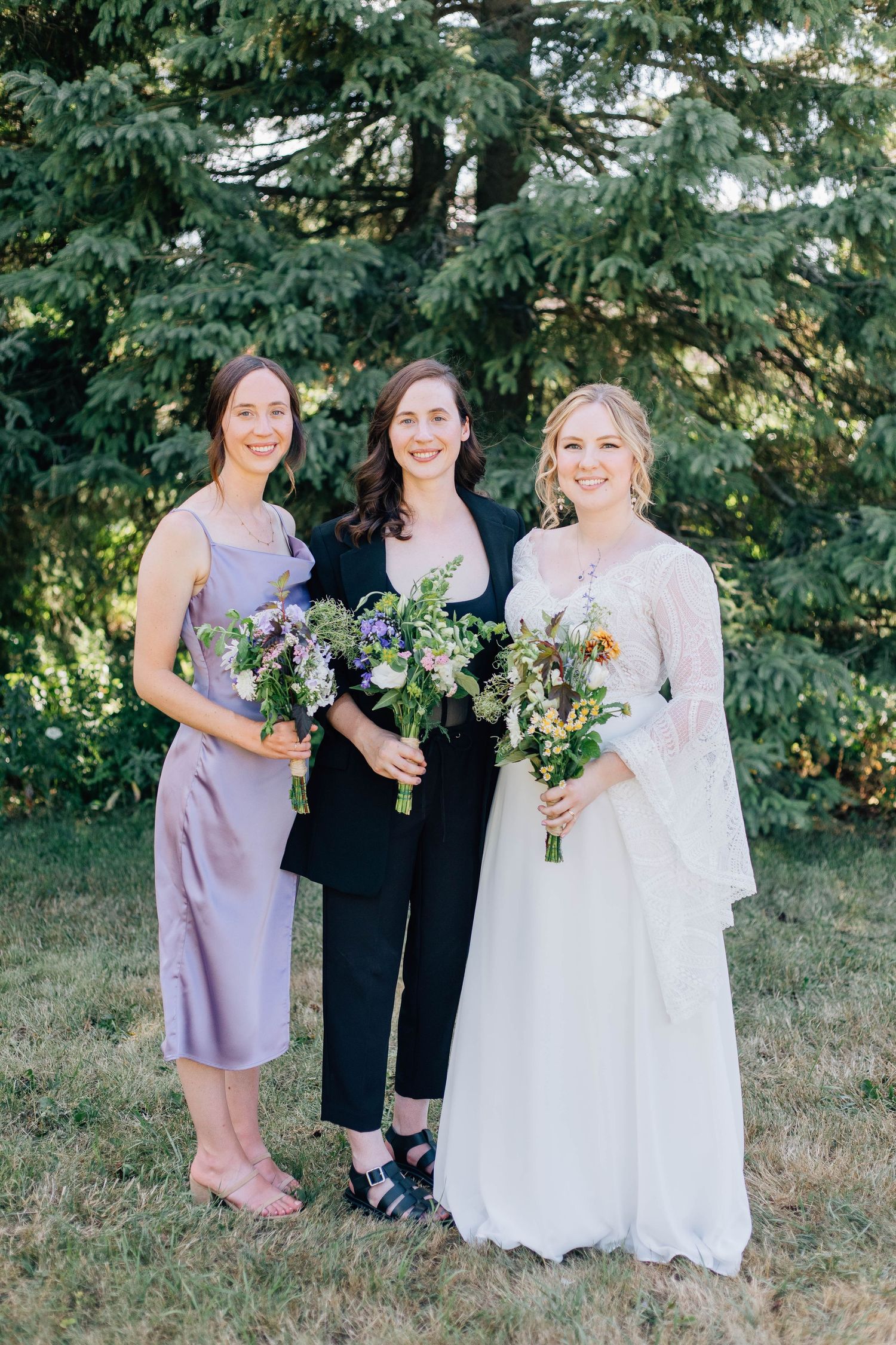Three women smile outdoors, bride in lace gown flanked by attendants holding wildflower bouquets.