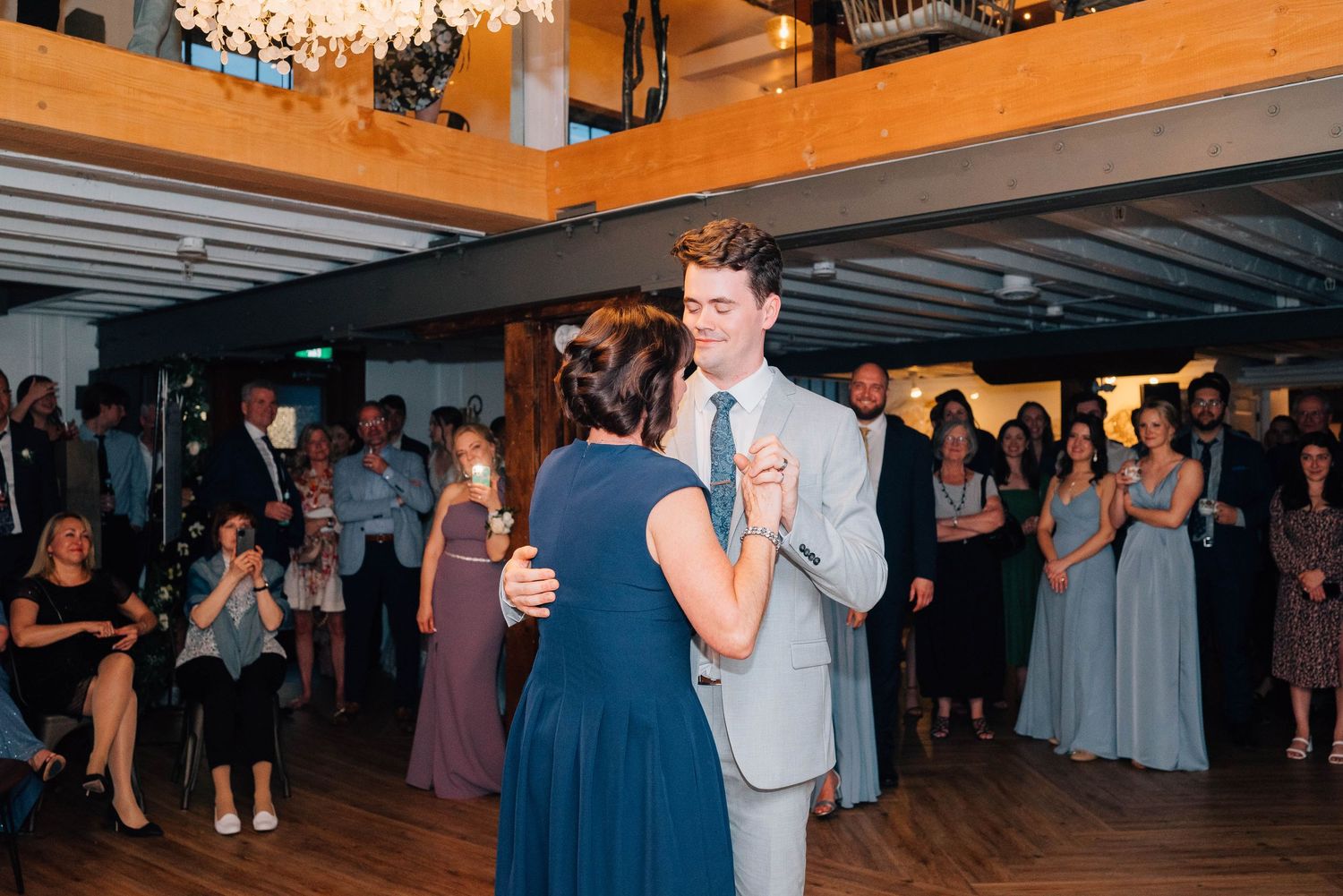 Groom in gray suit dances with a woman in blue dress at elegant wedding reception.