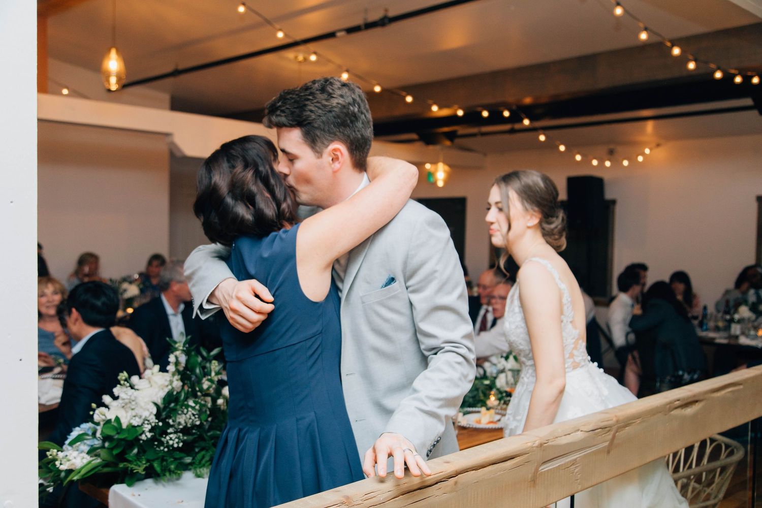 Groom in gray suit embracing mother in navy dress at wedding reception head table.