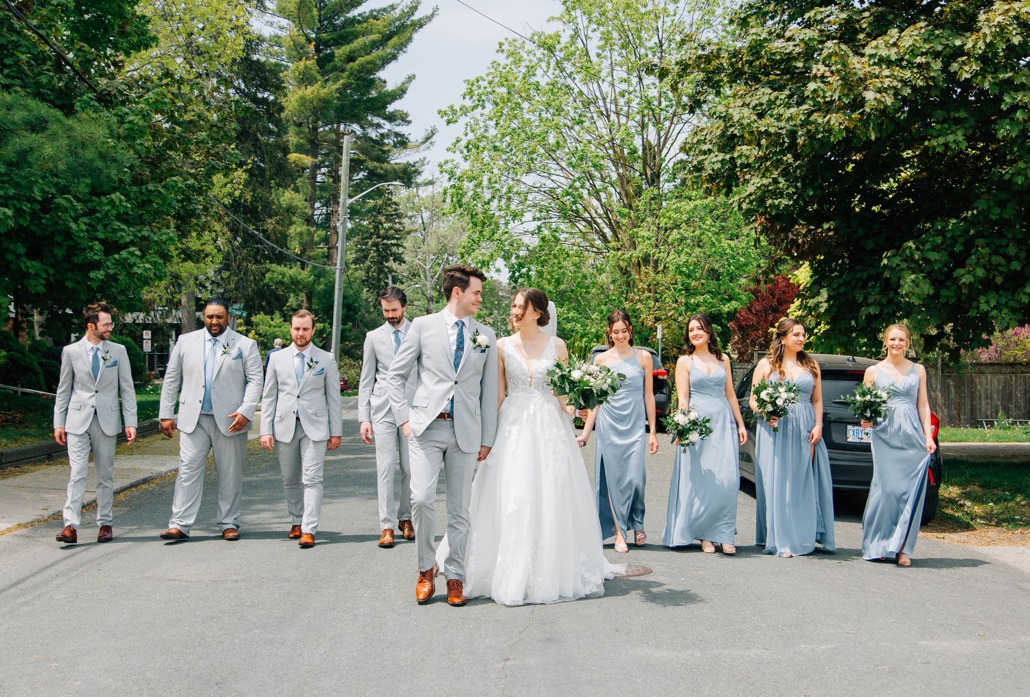 Wedding party walks together on a sunny road, groomsmen in gray suits and bridesmaids in light blue gowns.