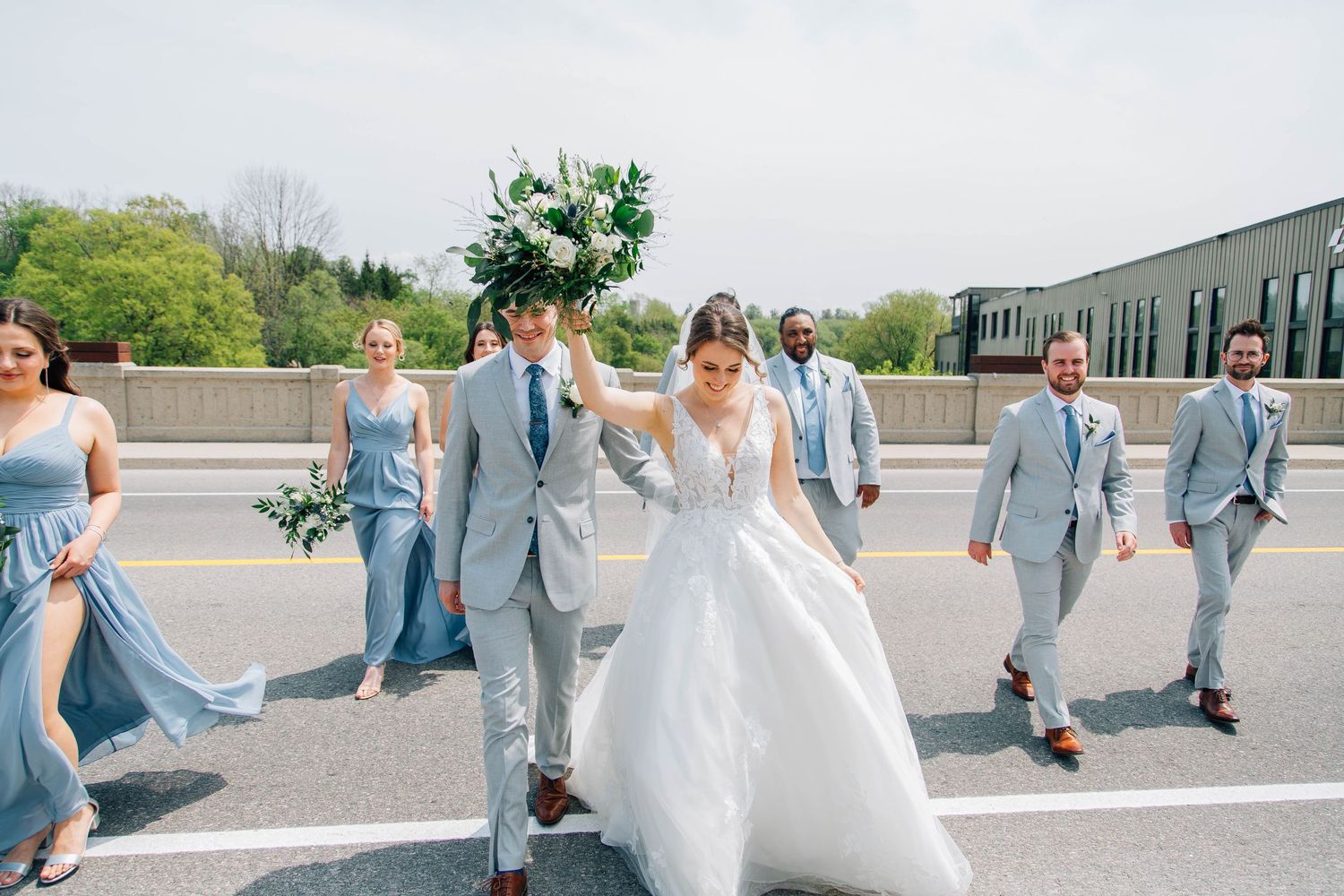 Bride and groom lead wedding party across bridge, bride holding lush greenery bouquet on sunny spring day.