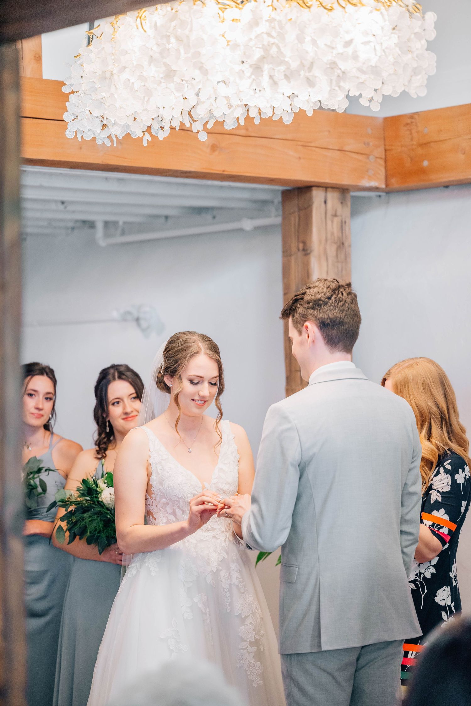 Bride placing wedding ring on groom's finger with bridesmaids in background.