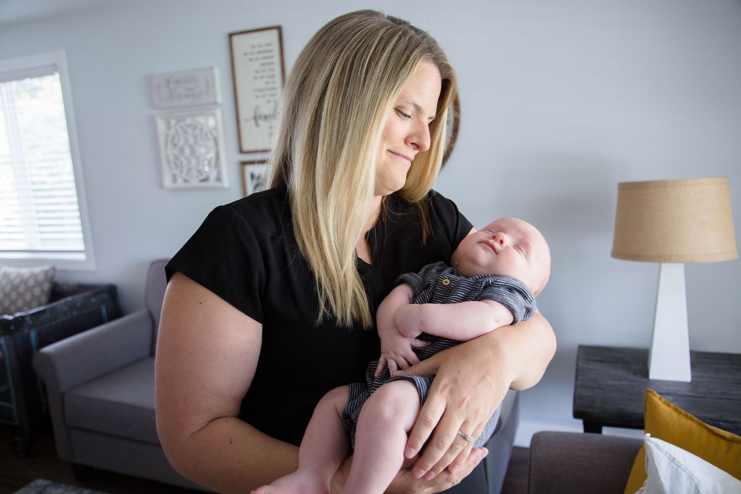 Blonde mother in black shirt holding newborn baby in gray onesie in bright home interior.