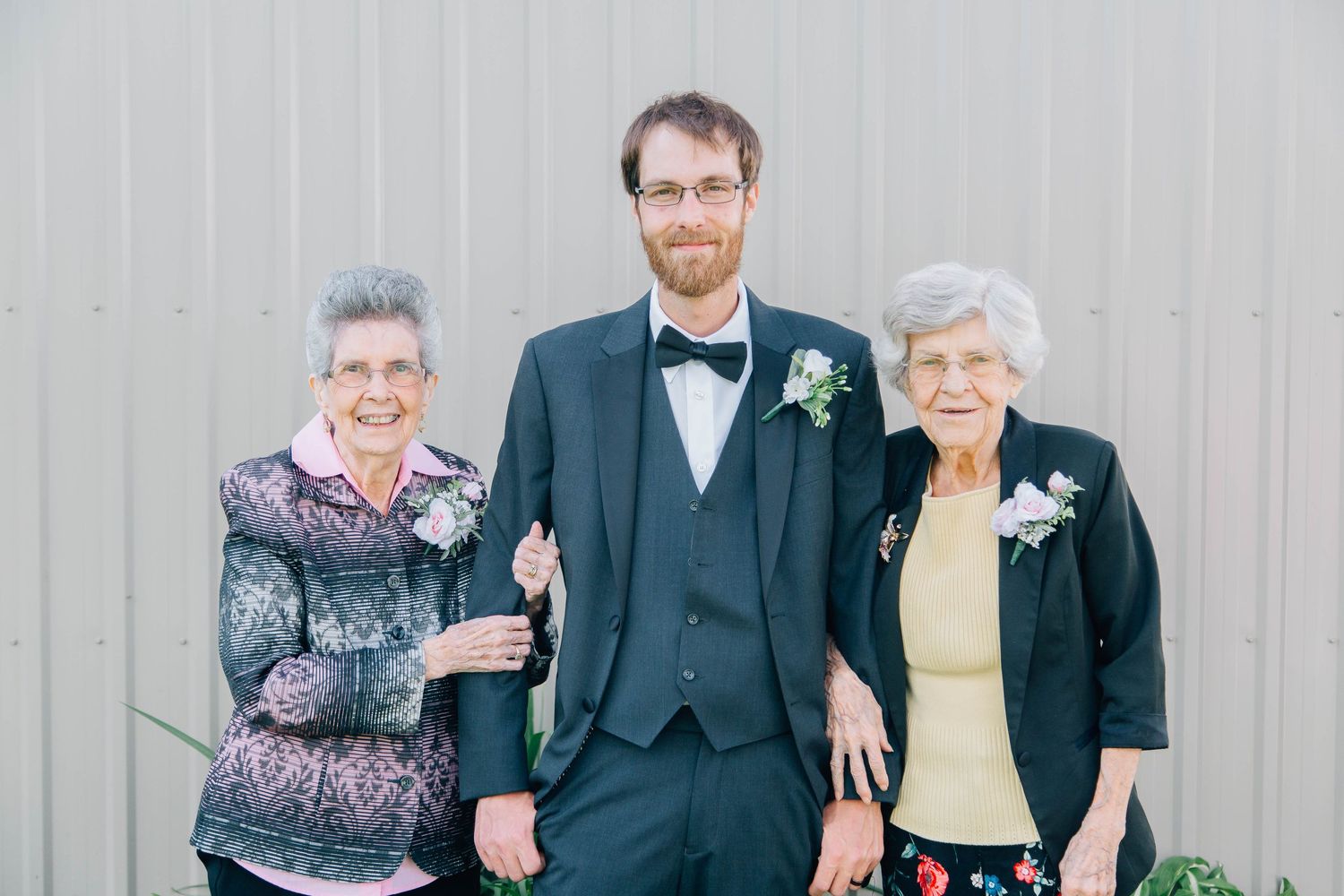 A groom in a teal suit poses with two elderly women wearing corsages outside a gray building.