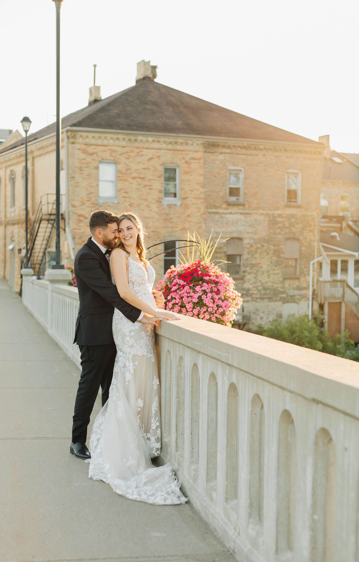 Bride and groom pose on a sunlit bridge with a vibrant pink floral bouquet and stone building backdrop in Paris Ontario.