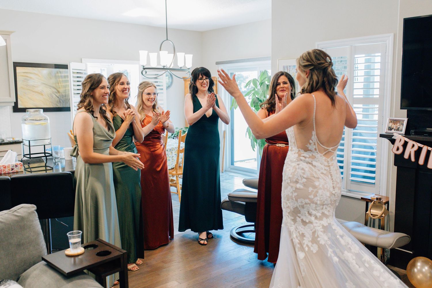 Bridesmaids react with joy and applause as bride in lace wedding gown reveals her look in a home living room.