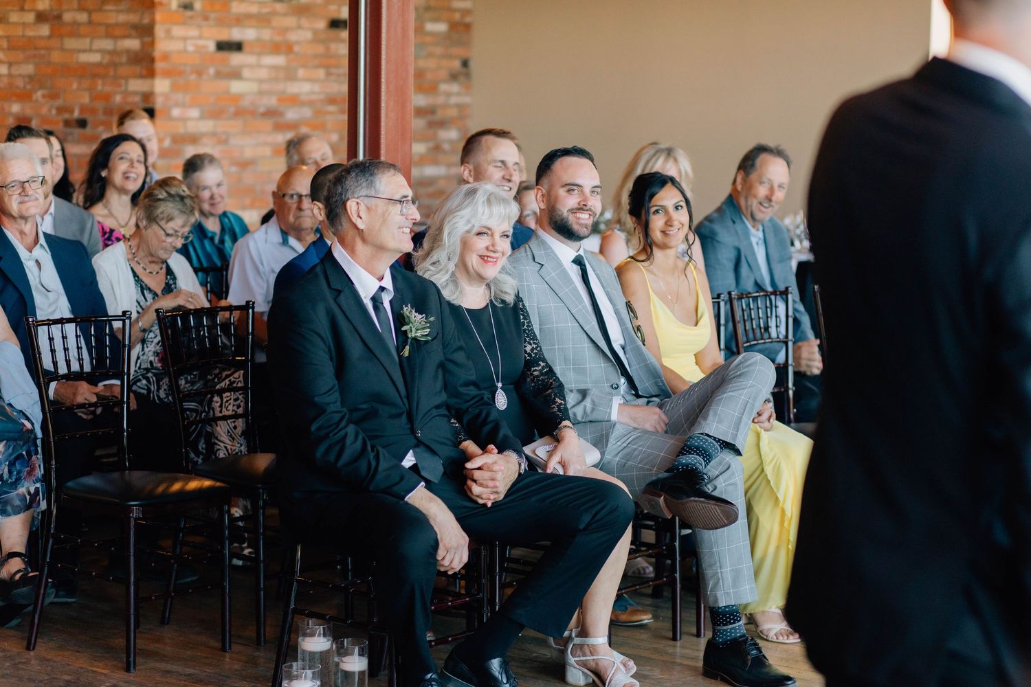 Smiling wedding guests seated in rows watching ceremony in bright indoor venue.