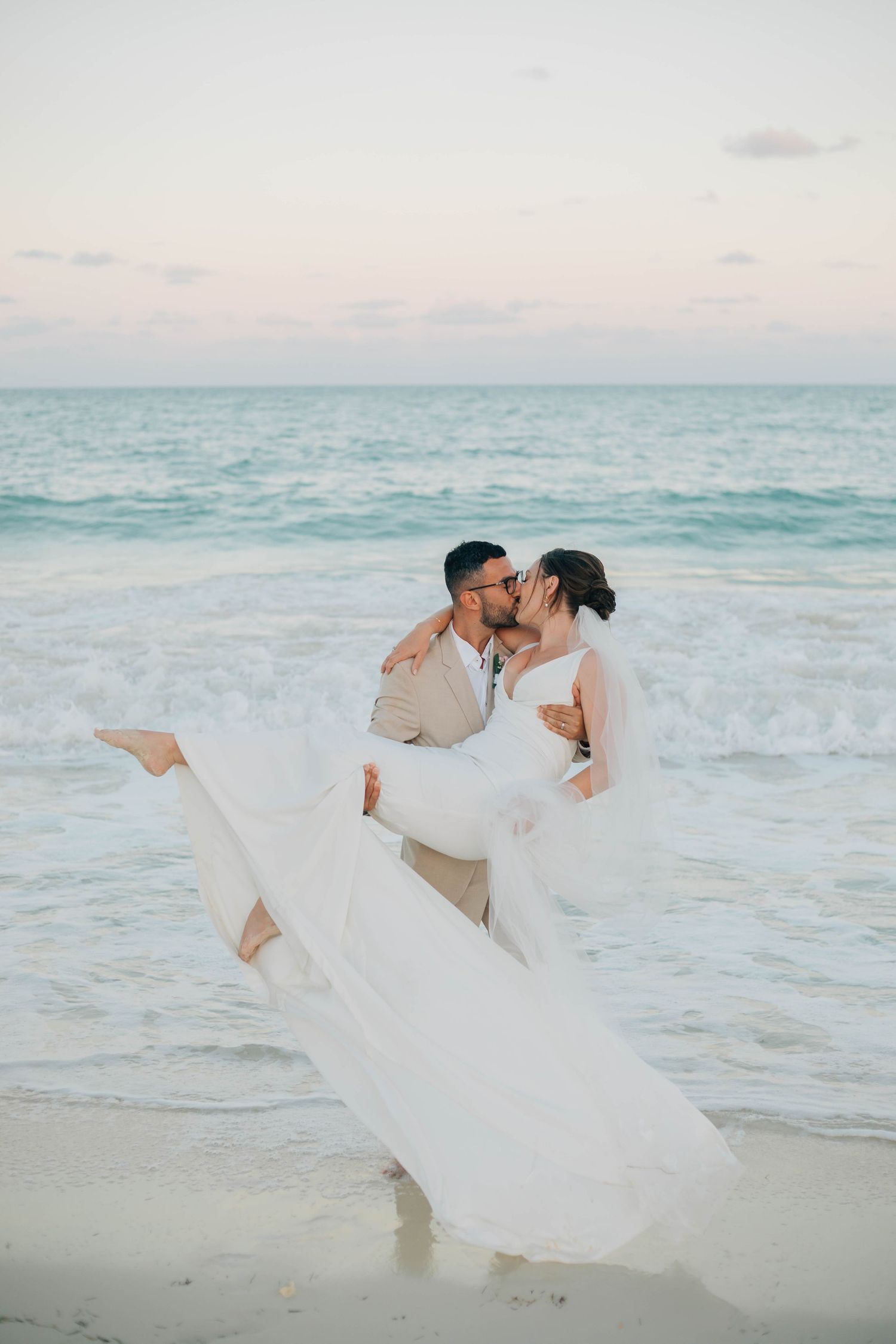Groom carries bride in white gown while kissing on a sandy beach at sunset with ocean waves behind them.