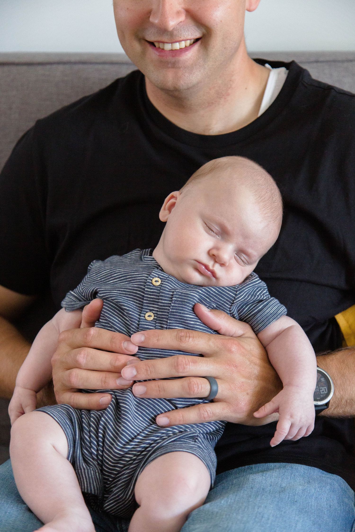 Smiling father in black t-shirt cradling sleeping newborn baby in striped onesie on a couch.