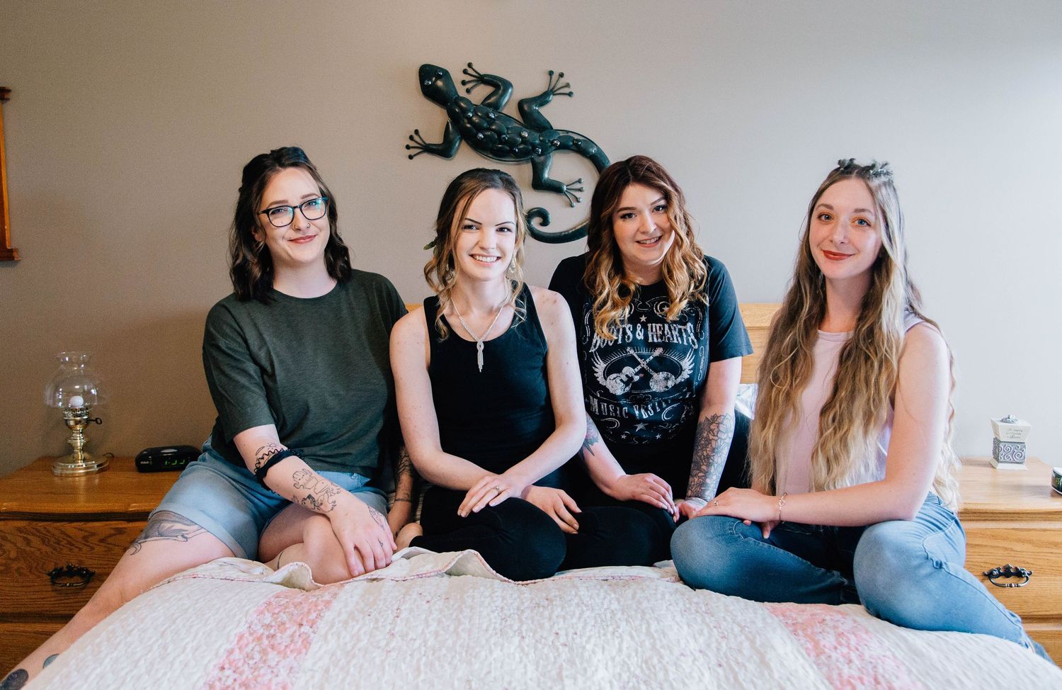 Four young women sitting together smiling on bed in rustic bedroom.