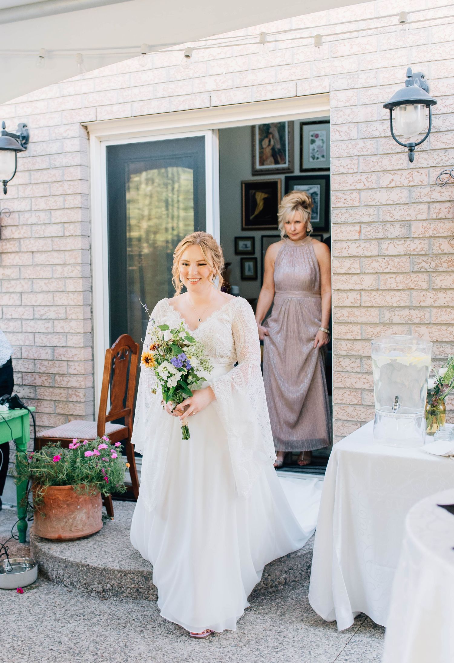 Blonde bride in white lace gown with long sleeves smiles while carrying colorful wildflower bouquet at backyard wedding.