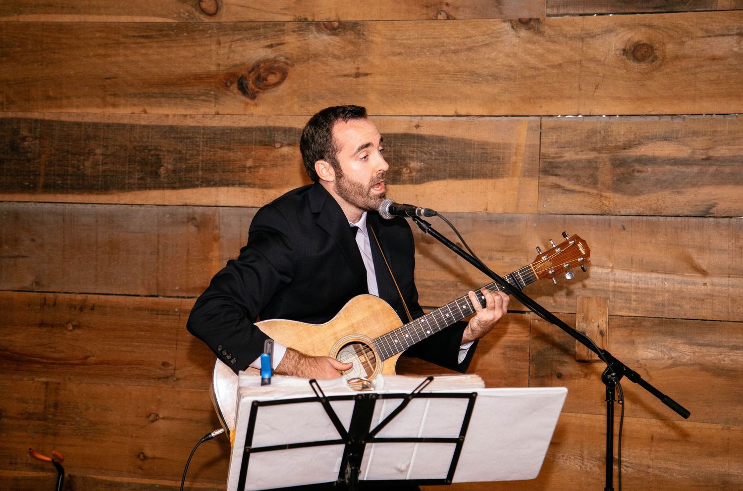 Live musician playing acoustic guitar and singing at a wedding reception in a rustic barn.