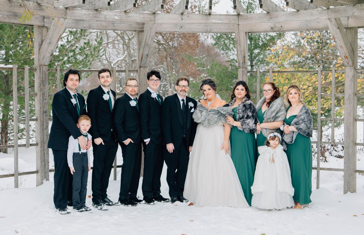Full winter wedding party poses in snow under pergola in coordinating emerald green and black attire.