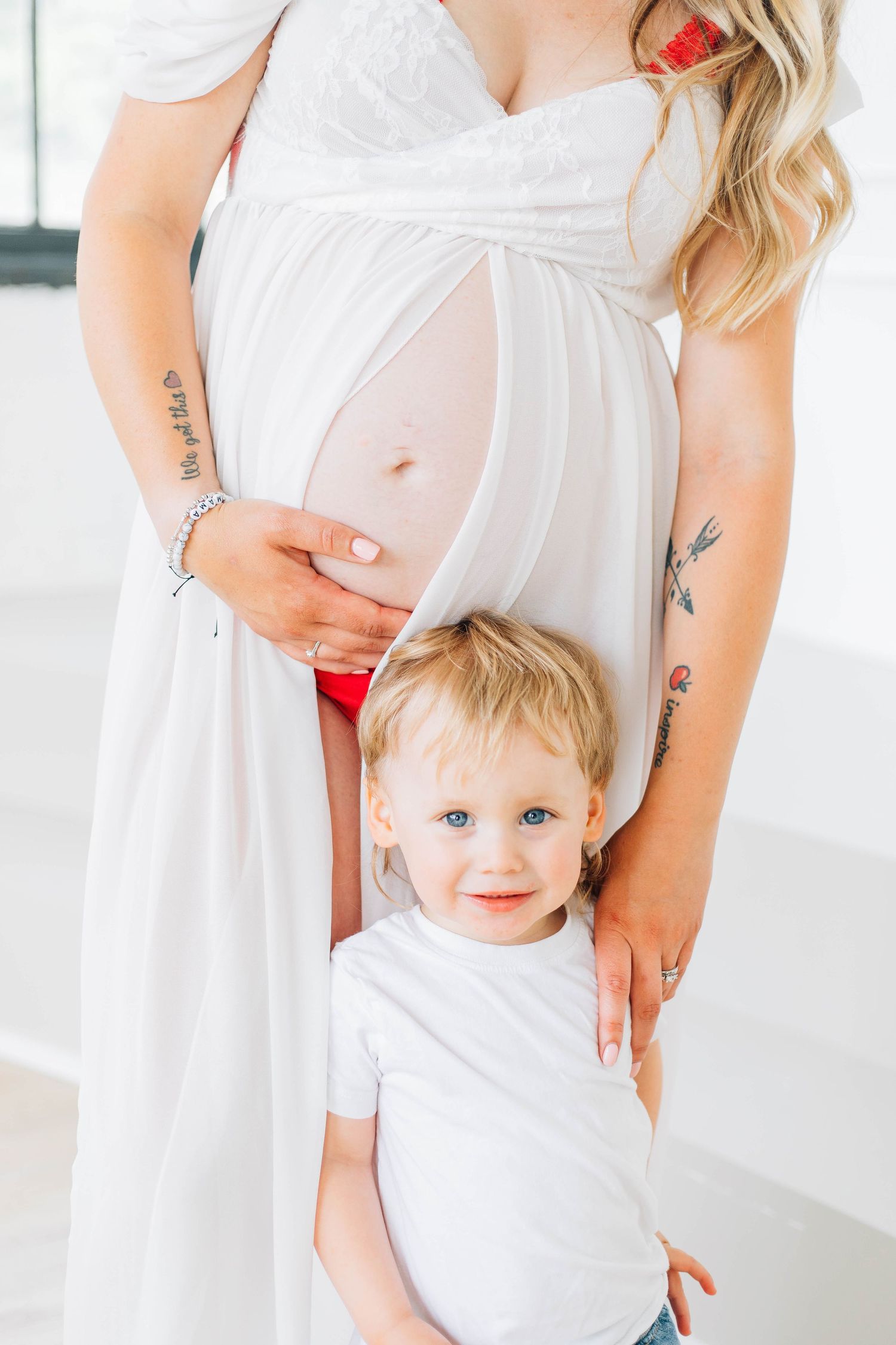 Smiling toddler standing in front of pregnant mother in sheer white maternity gown.